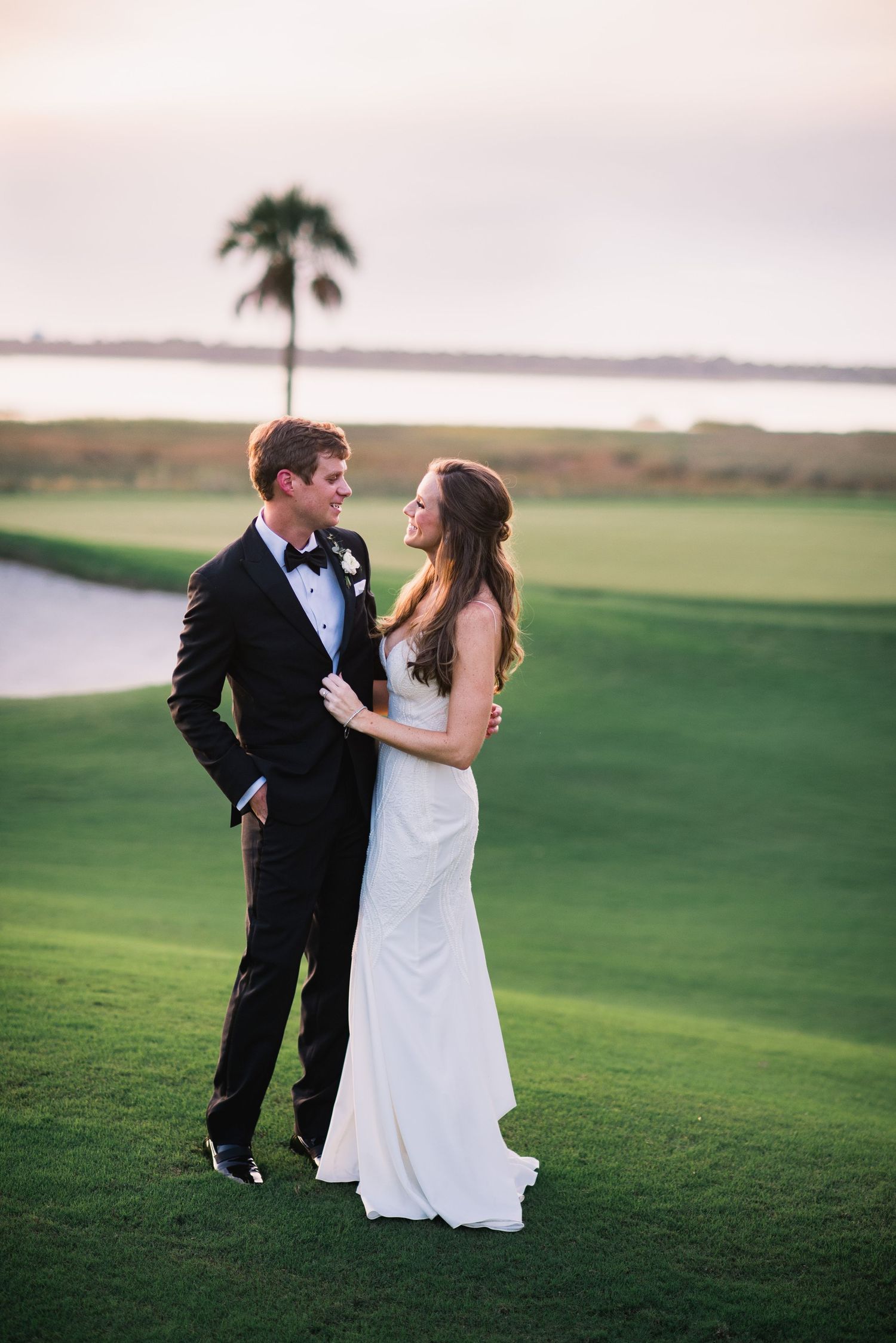 A couple in wedding attire embraces on a green lawn overlooking a waterfront during golden hour.