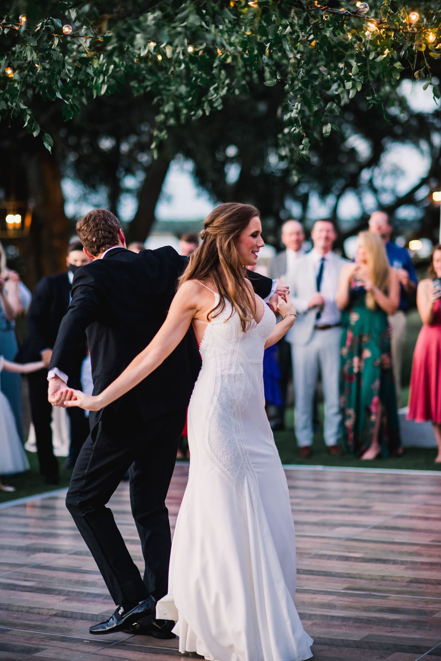 A couple dances together at an outdoor wedding reception on a wooden deck as guests watch in the background.