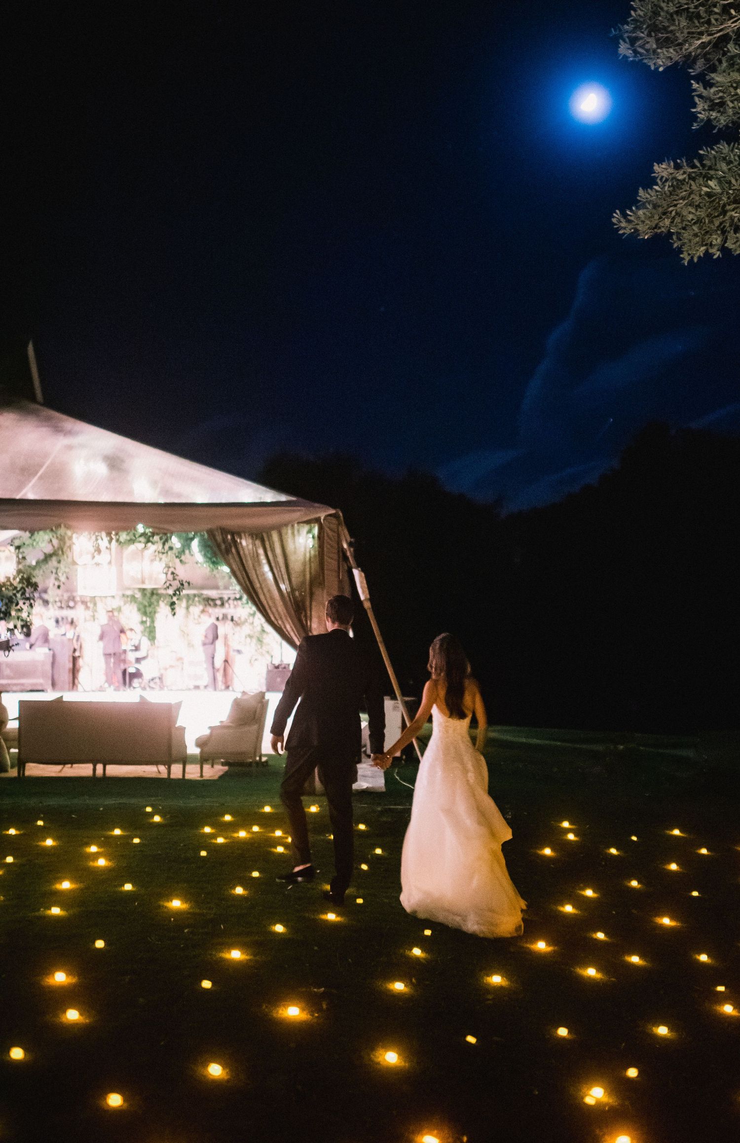 A romantic outdoor wedding reception at night with illuminated lanterns lining the path to a glowing white tent.