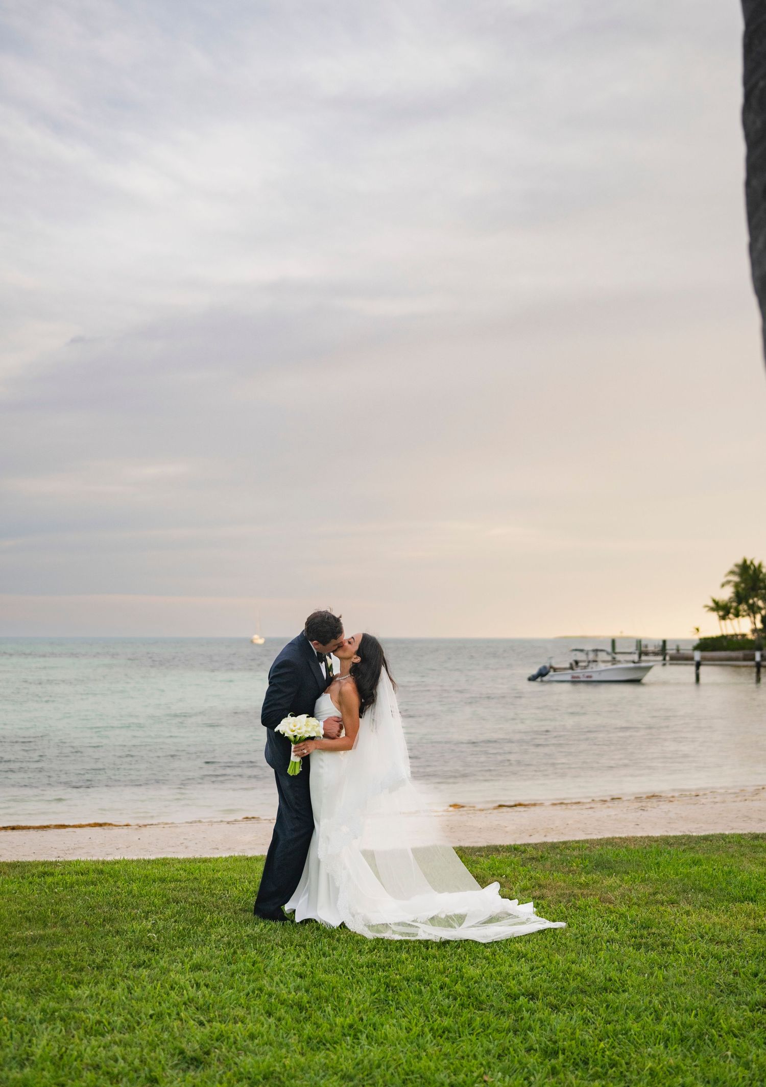 A romantic wedding couple embraces on a scenic beach with green grass and ocean views at sunset.