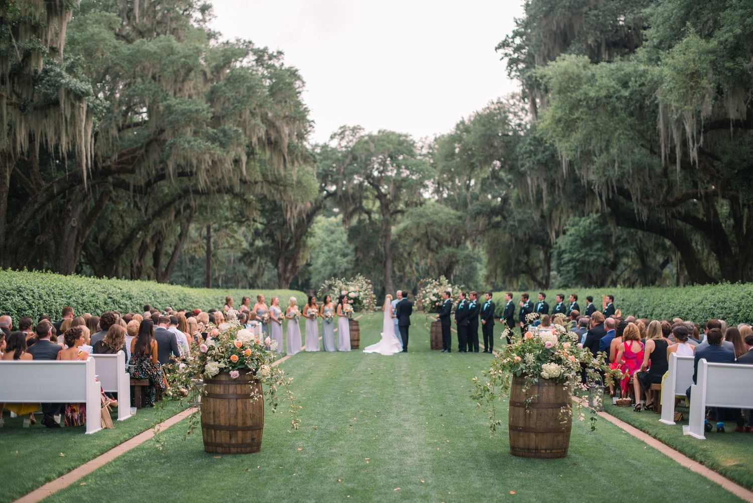 Guests gather for an outdoor wedding ceremony under Spanish moss-draped oak trees on a lush green lawn.