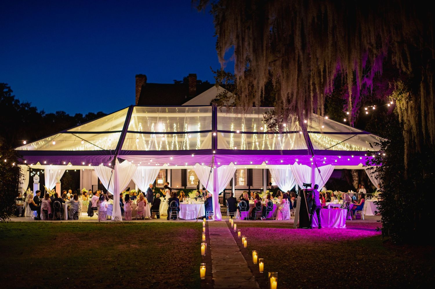 Outdoor wedding reception tent illuminated with purple uplighting at night creating an elegant atmosphere.