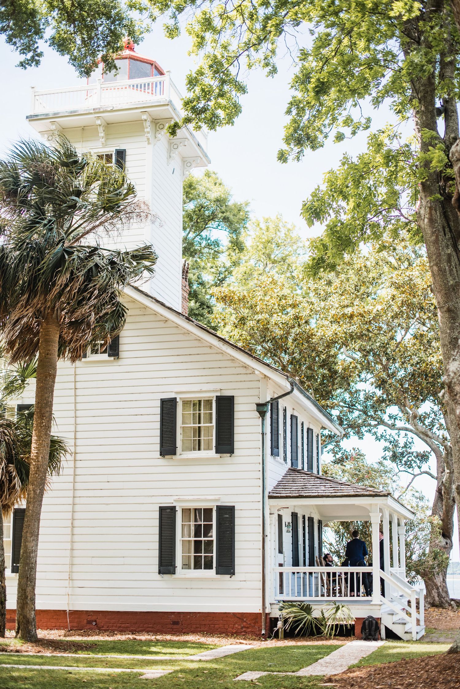 Historic white lighthouse keeper's house with black shutters and wraparound porch nestled among oak trees and palm trees.