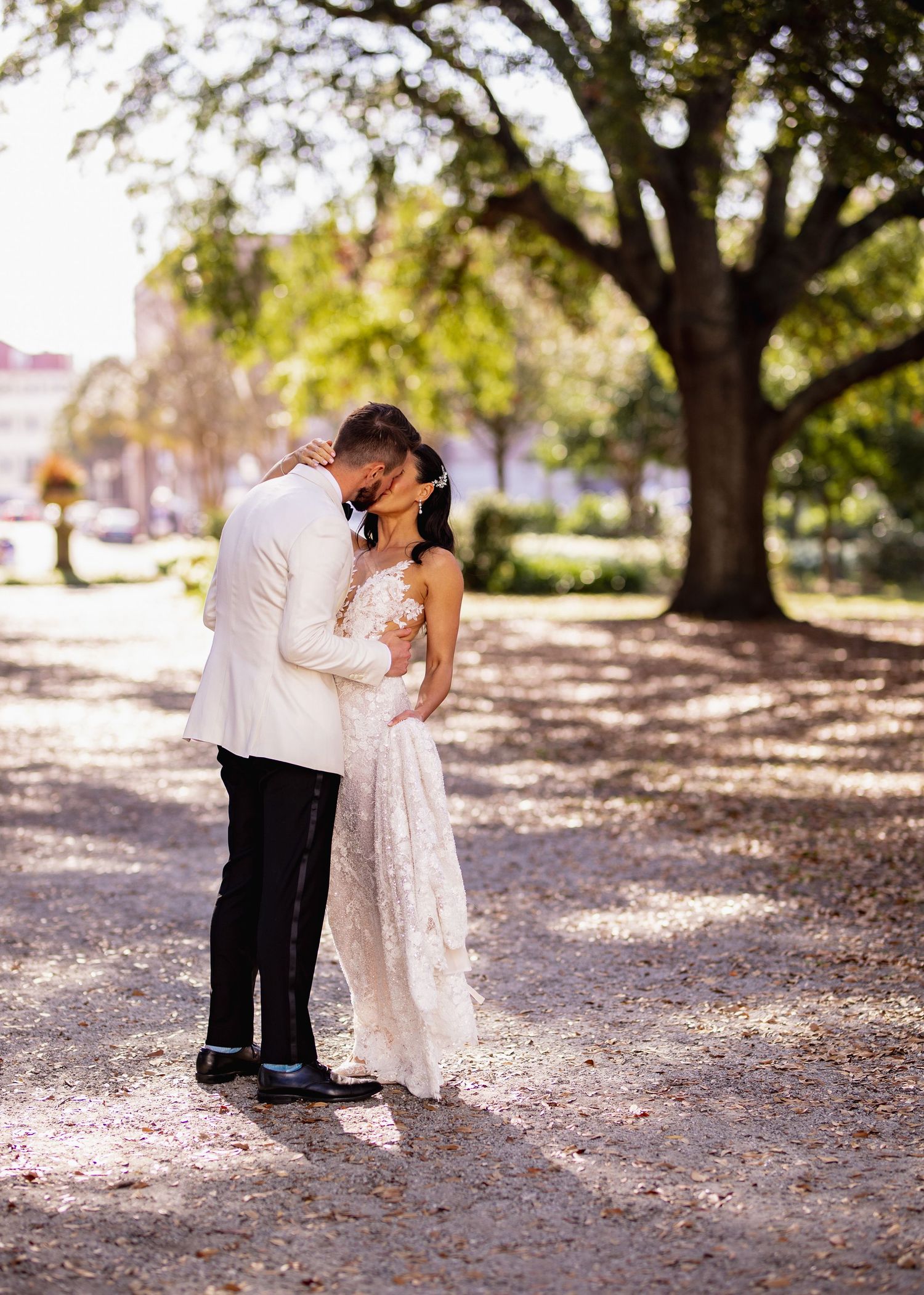 A couple in wedding attire share an intimate moment under oak trees with autumn leaves scattered on the ground.