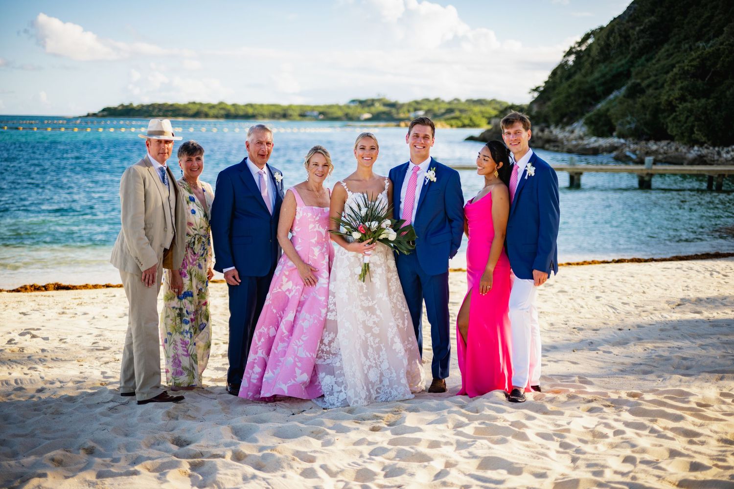Wedding party poses for group photos on a tropical beach with blue ocean and green hills in background.