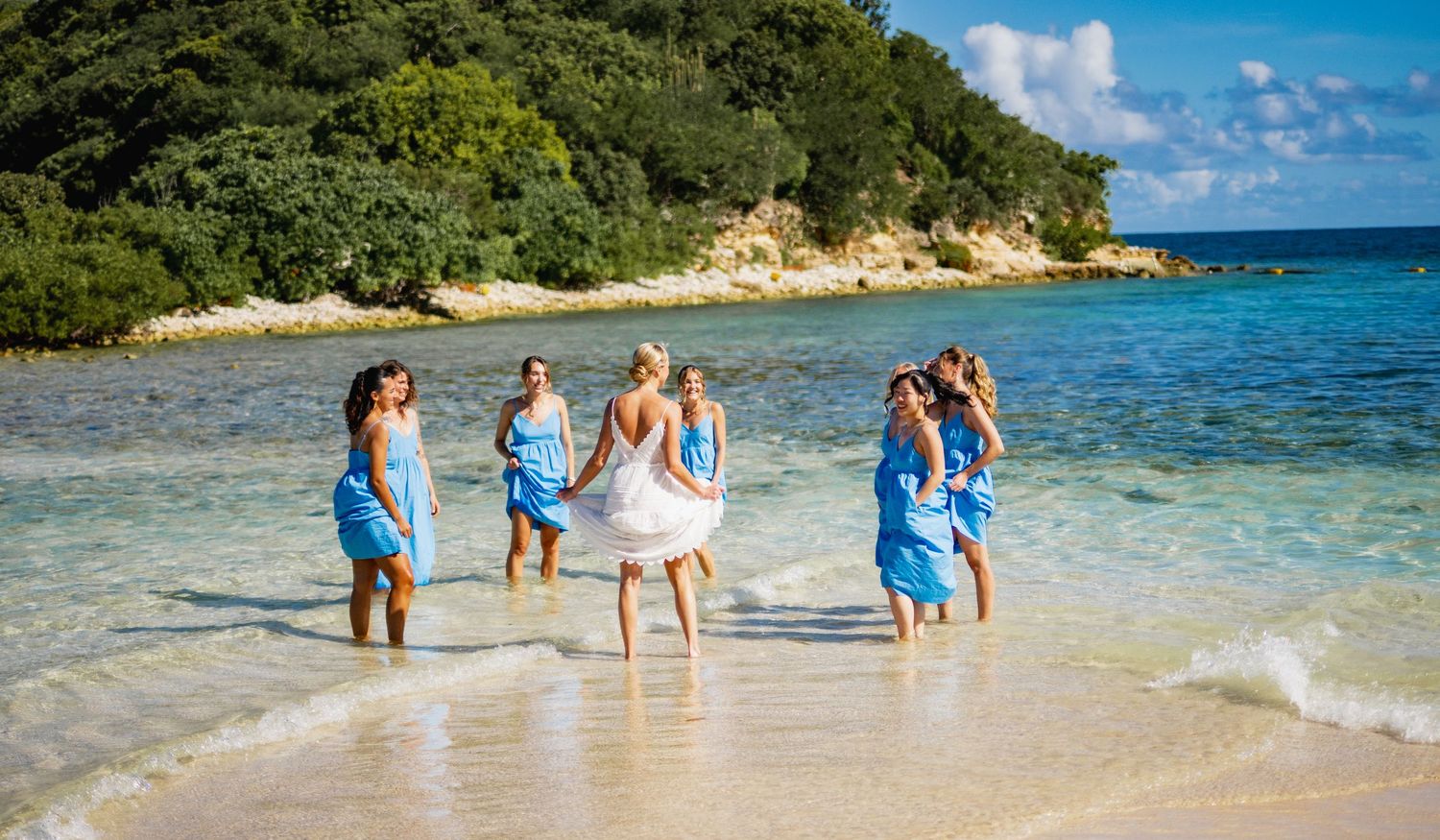 A group in blue dresses and white dress enjoying a sunny day at a tropical beach with clear turquoise waters.