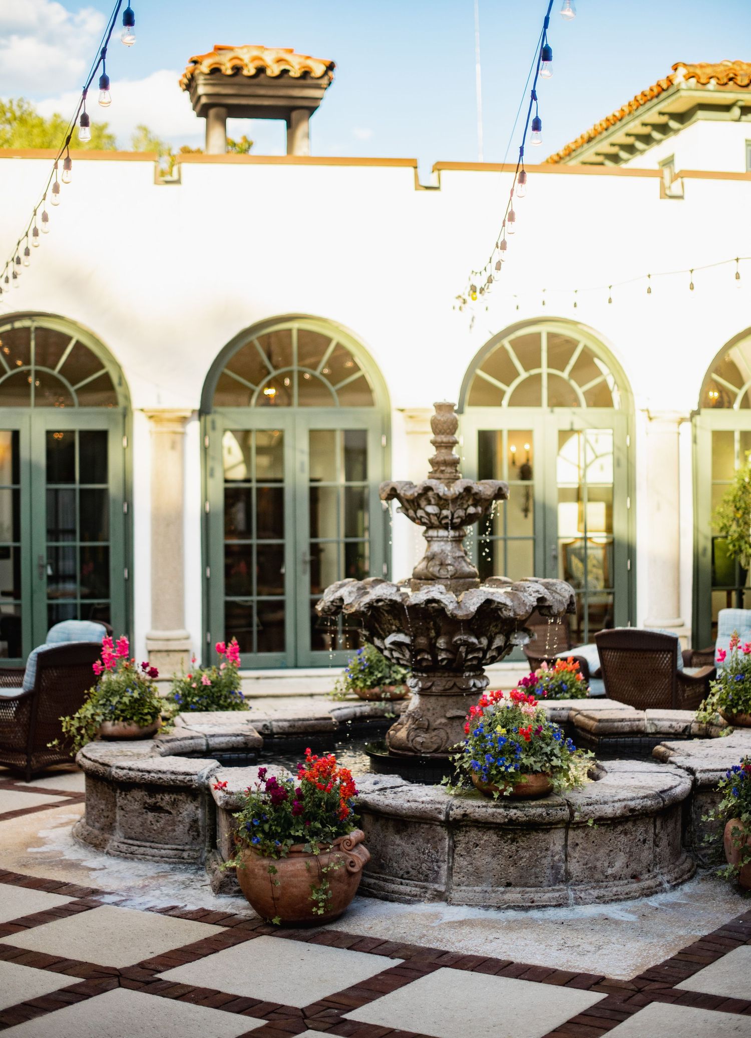 A curved stone garden wall with pink flowers and decorative urns lines a Mediterranean-style courtyard with arched windows.
