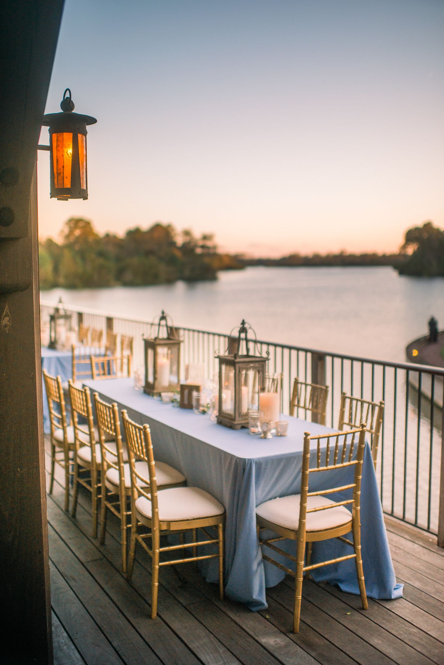 Elegant outdoor dining setup with gold chiavari chairs and light blue tablecloths on a lakeside deck at sunset.