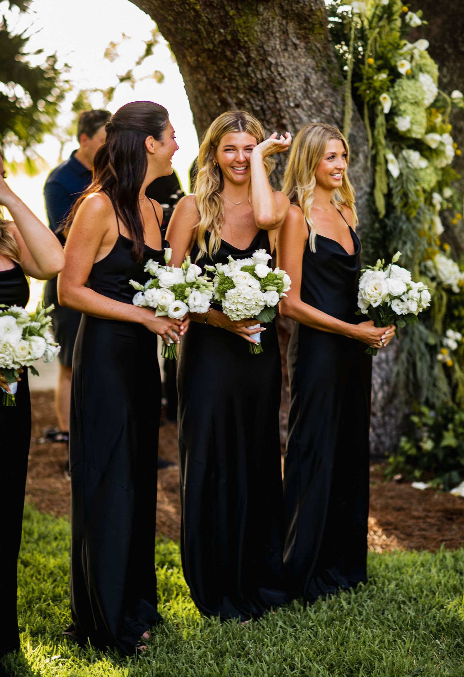 A line of bridesmaids in elegant black dresses holding white bouquets stand together on green grass during an outdoor wedding.