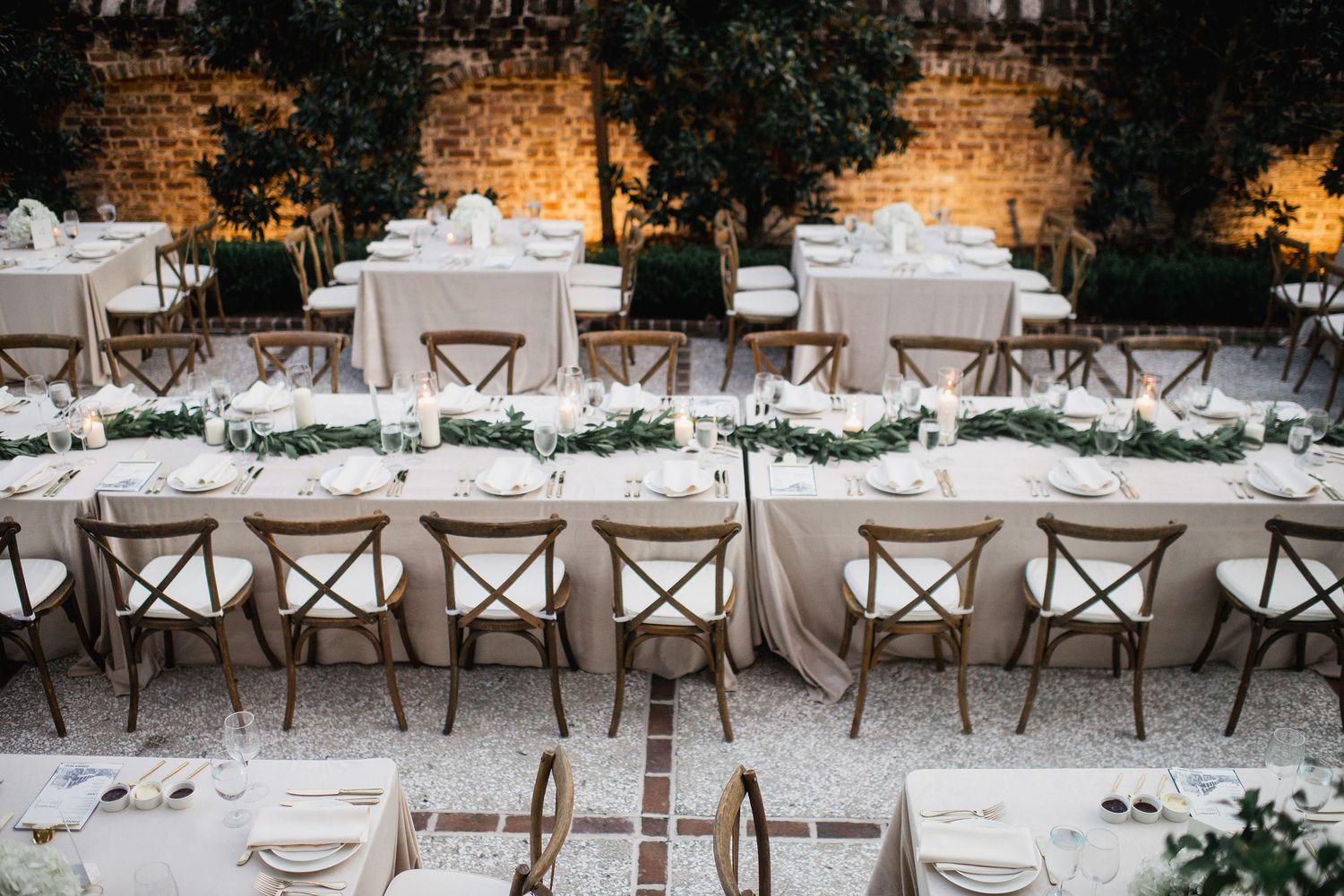 Elegant outdoor wedding reception setup with long tables, wooden cross-back chairs, and white linens against stone walls.