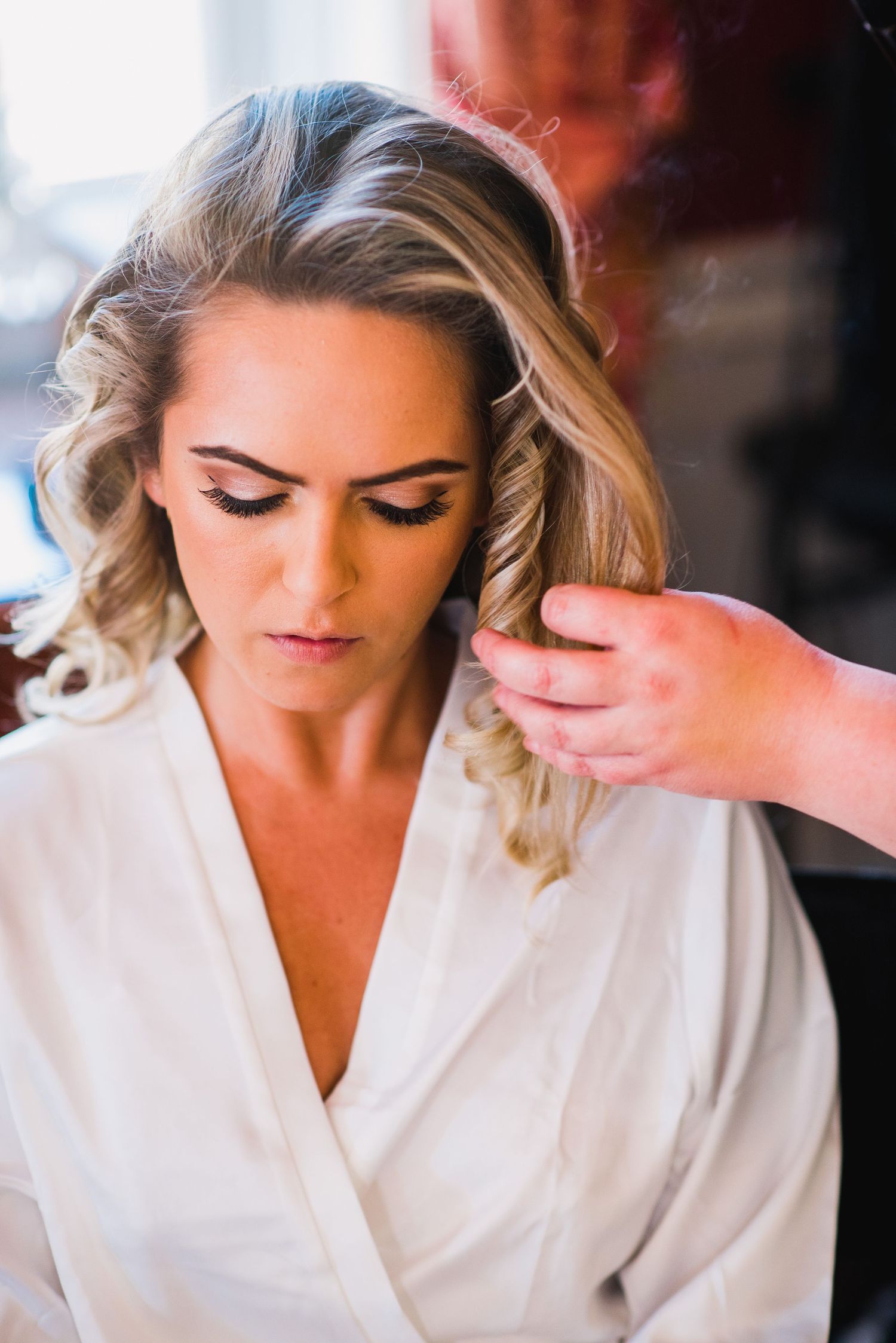 Professional makeup artist applies finishing touches to a bride wearing a white silk robe during wedding preparations.