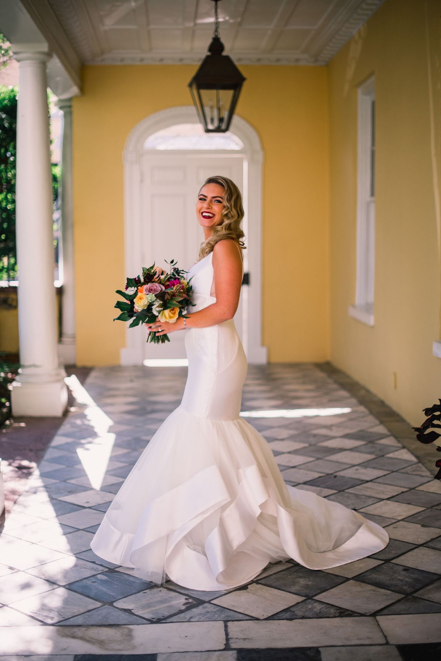 A bride in a white mermaid wedding gown poses on a tiled porch with yellow walls and white columns.