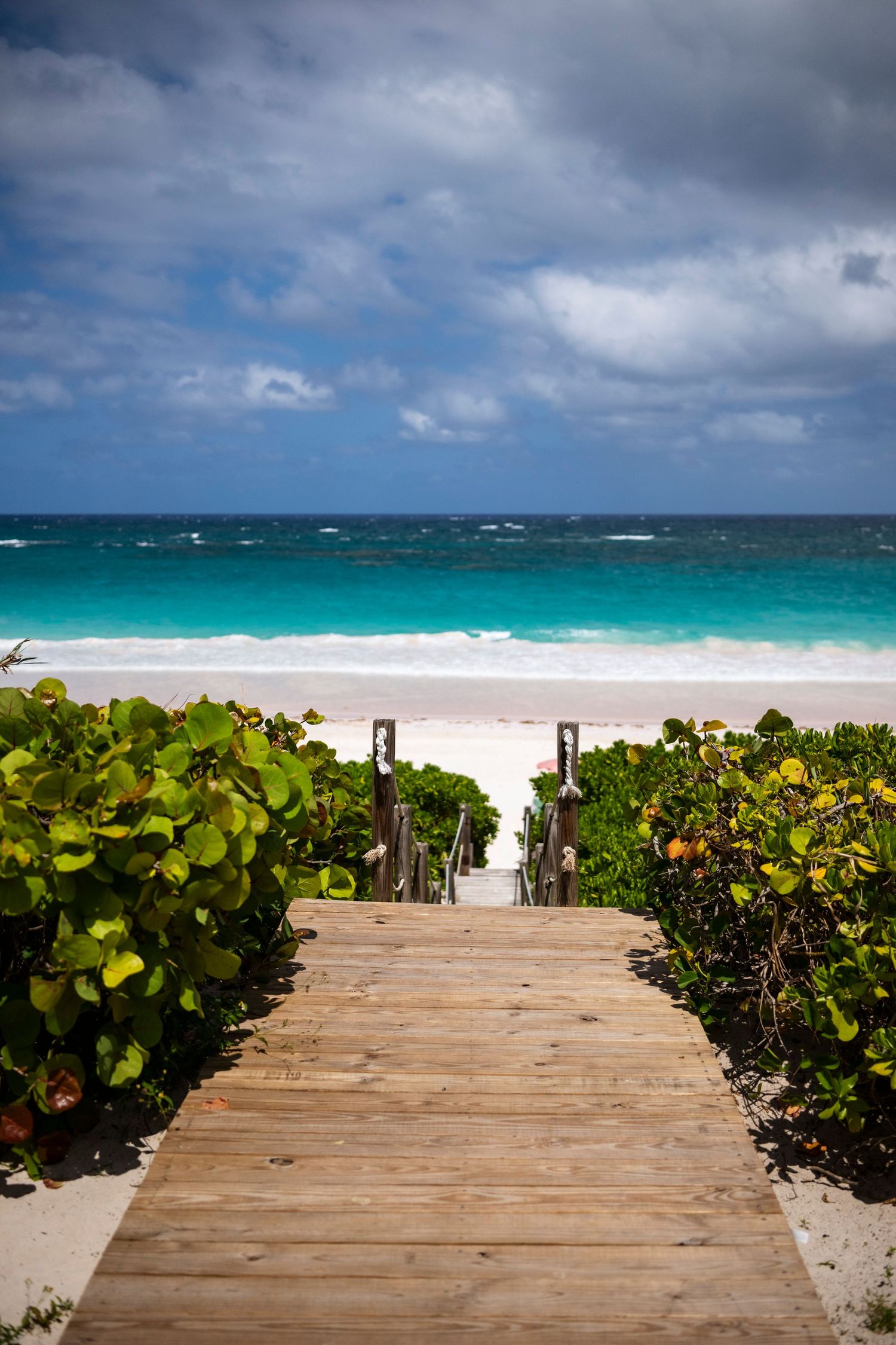 Wooden boardwalk path leading to a tropical beach with turquoise water and cloudy skies. on harbour island Bahamas
