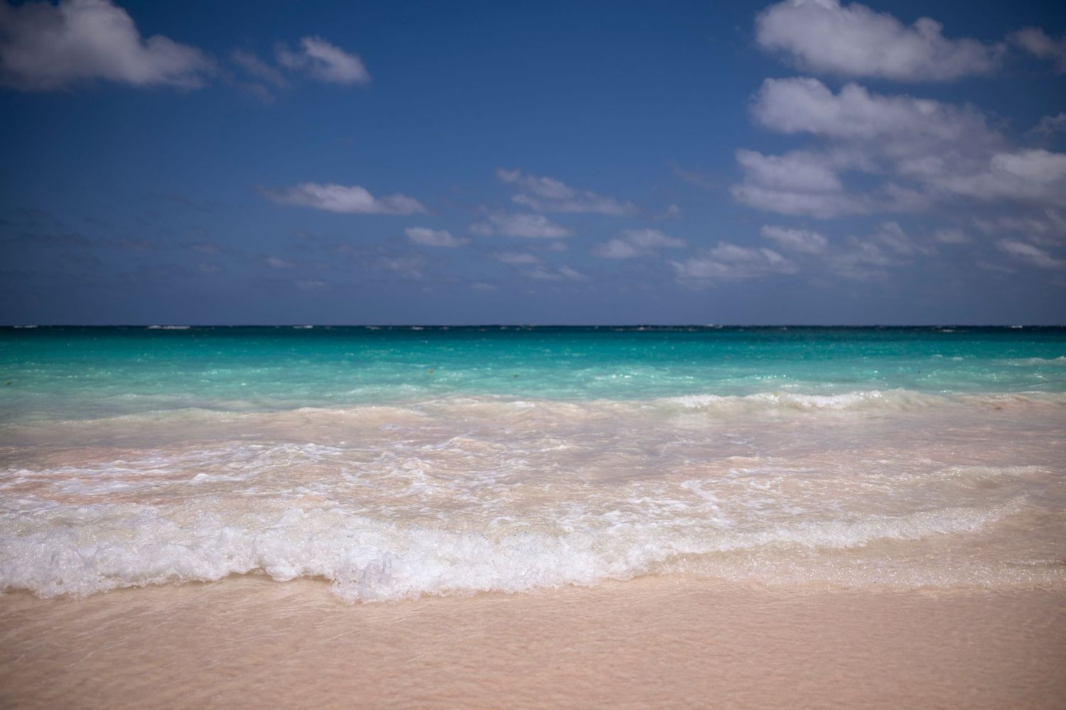 Pristine turquoise ocean waves meet white sandy beach under blue sky with scattered clouds.