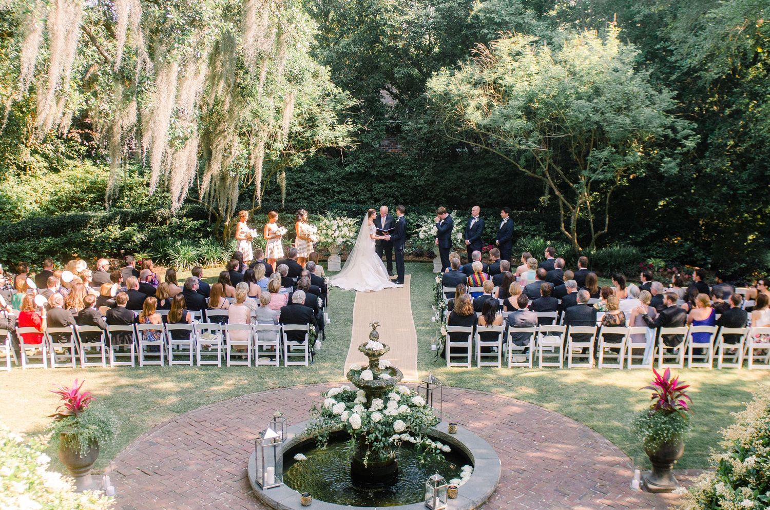 Outdoor garden wedding ceremony setup with rows of white chairs and floral arrangements along a tree-lined pathway.