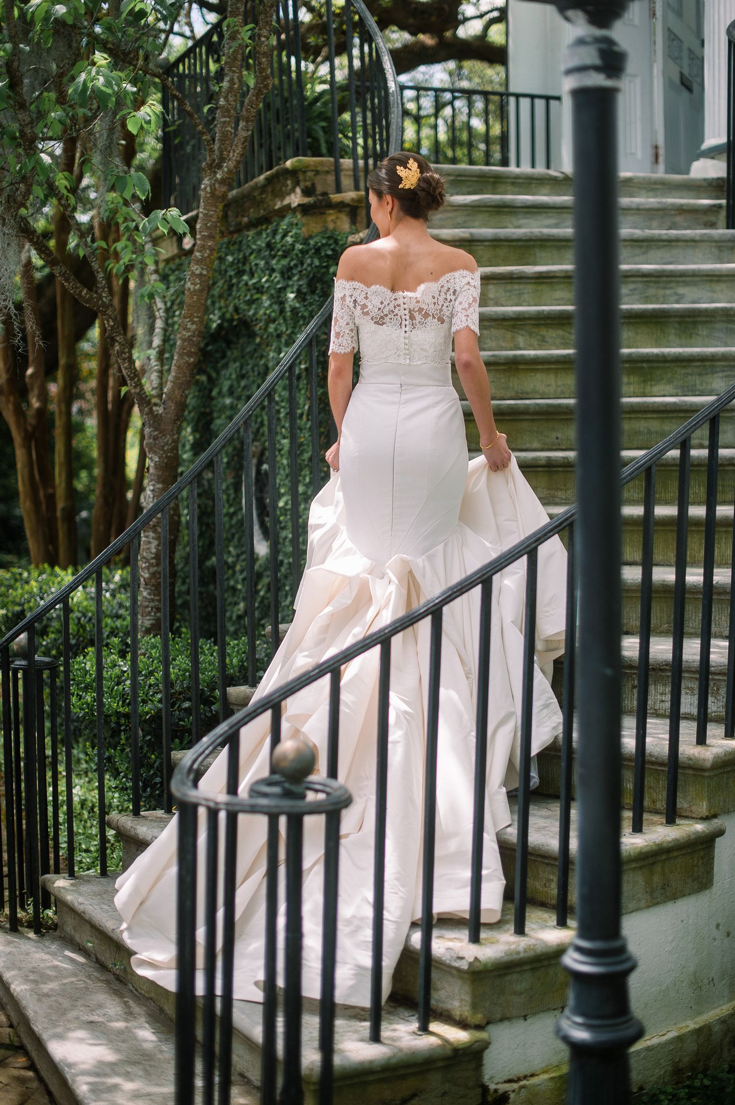 A bride in a white lace gown gracefully descends stone steps with an ornate black iron railing in a garden setting.