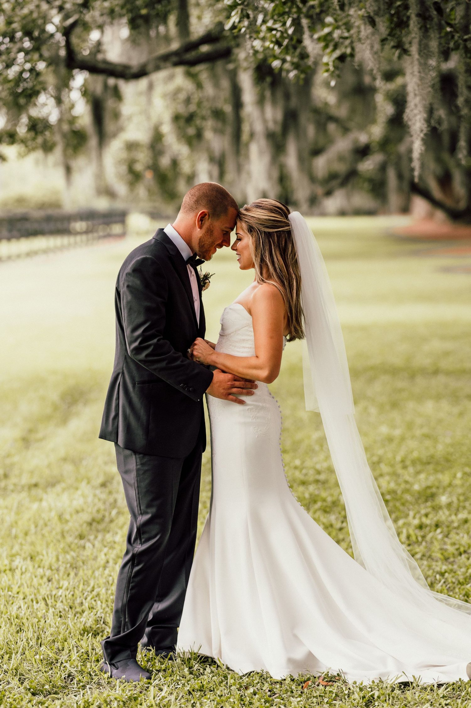 A romantic wedding couple shares an intimate moment under moss-draped oak trees in a sunlit garden setting.