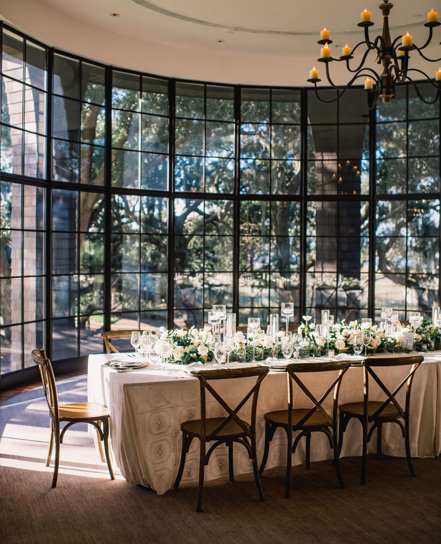Elegant wedding reception venue with floor-to-ceiling windows, rustic cross-back chairs, and white floral centerpieces.