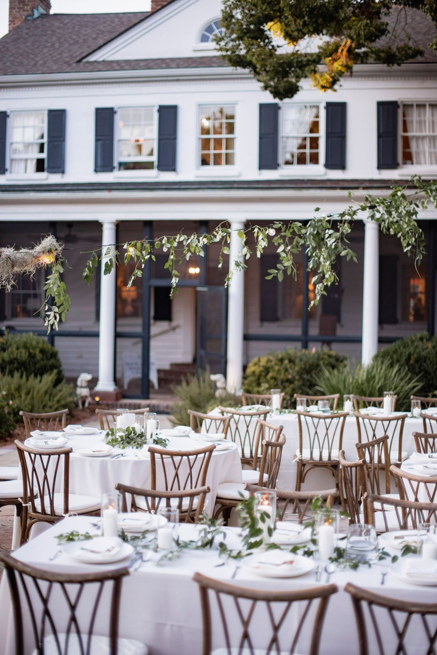 Elegant outdoor wedding reception setup with white-clothed tables and wooden chairs on a patio with colonial columns.