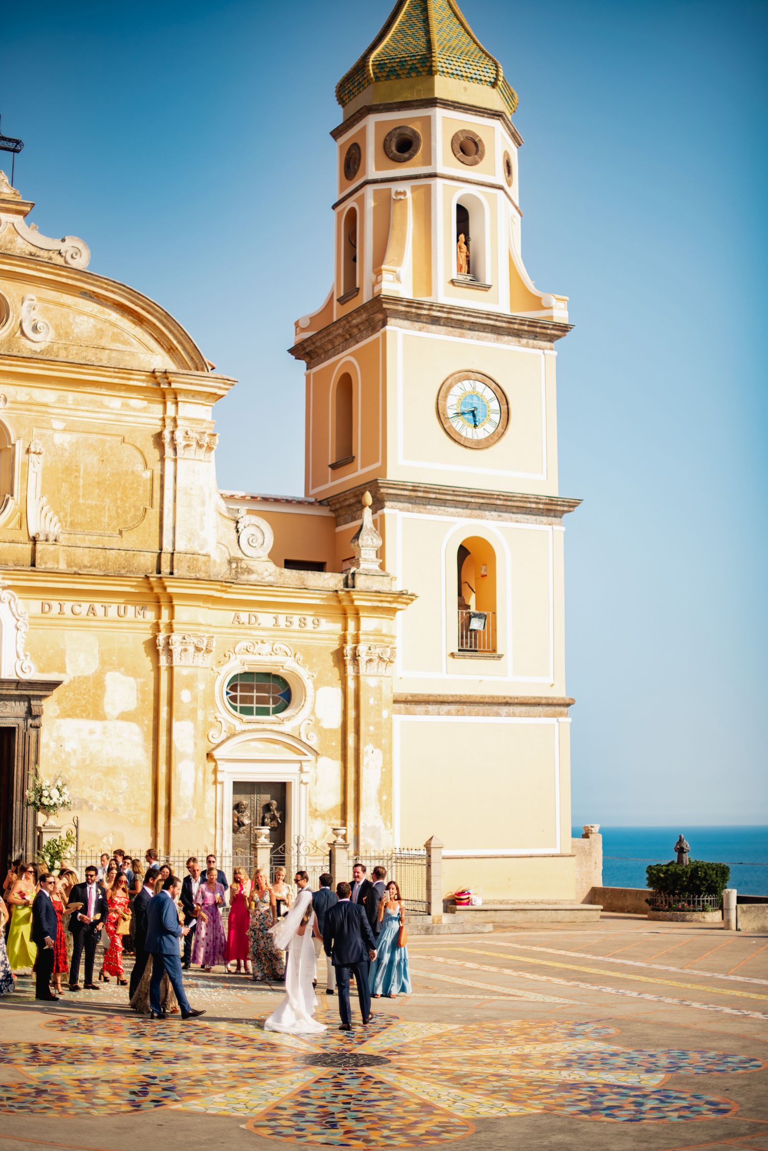 Wedding ceremony taking place at a Mediterranean church with yellow facade and ornate architecture overlooking the sea.