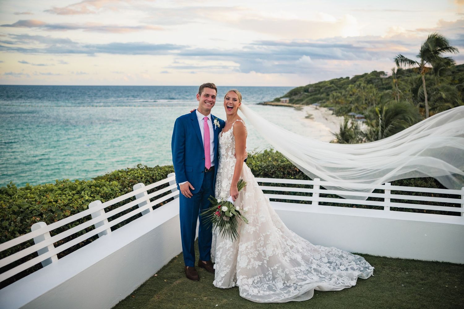 A couple shares romantic moments on a scenic beach with white railings and tropical views in the background.