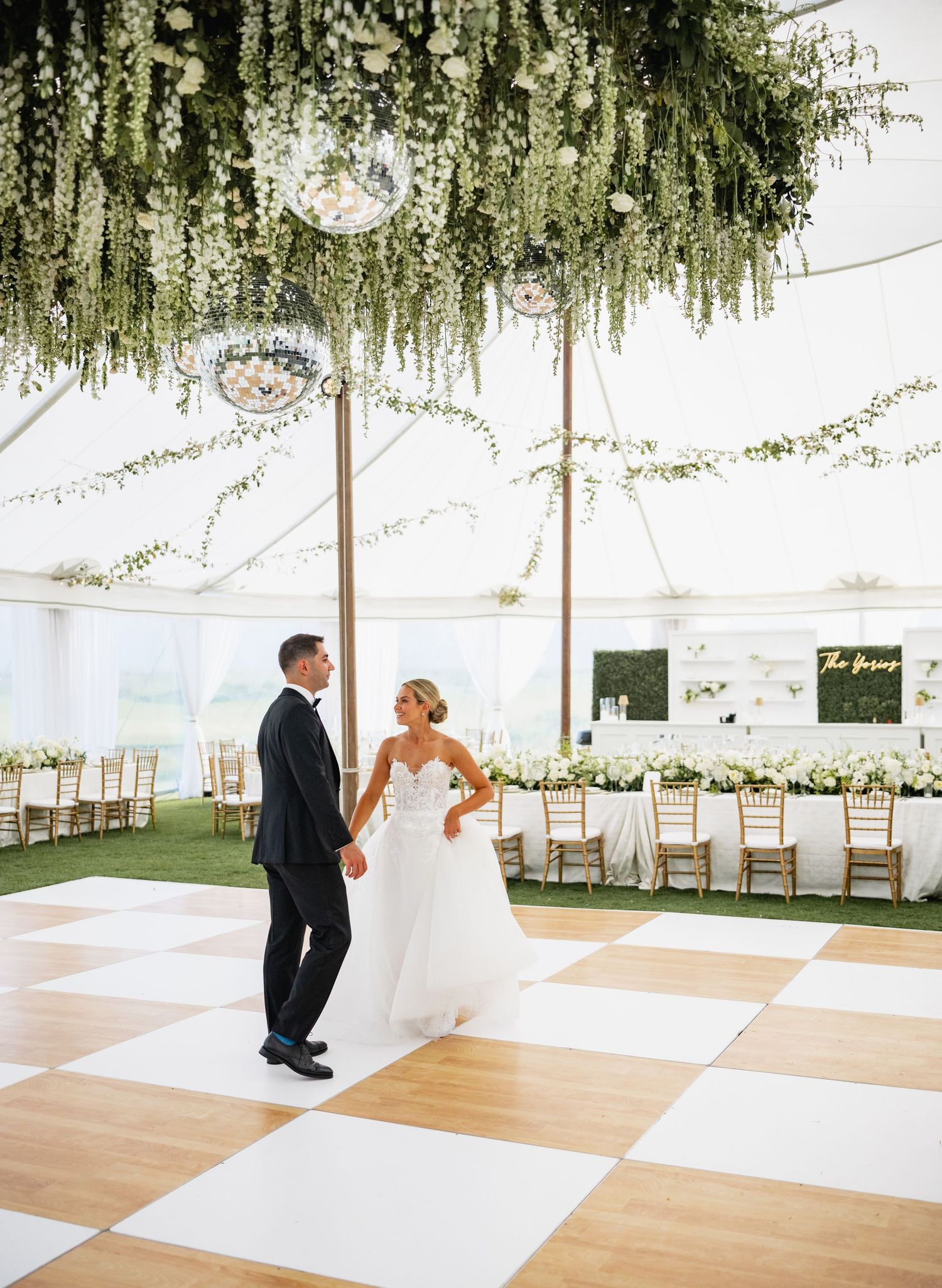 Elegant wedding reception space with checkered dance floor under hanging greenery and white tent ceiling.