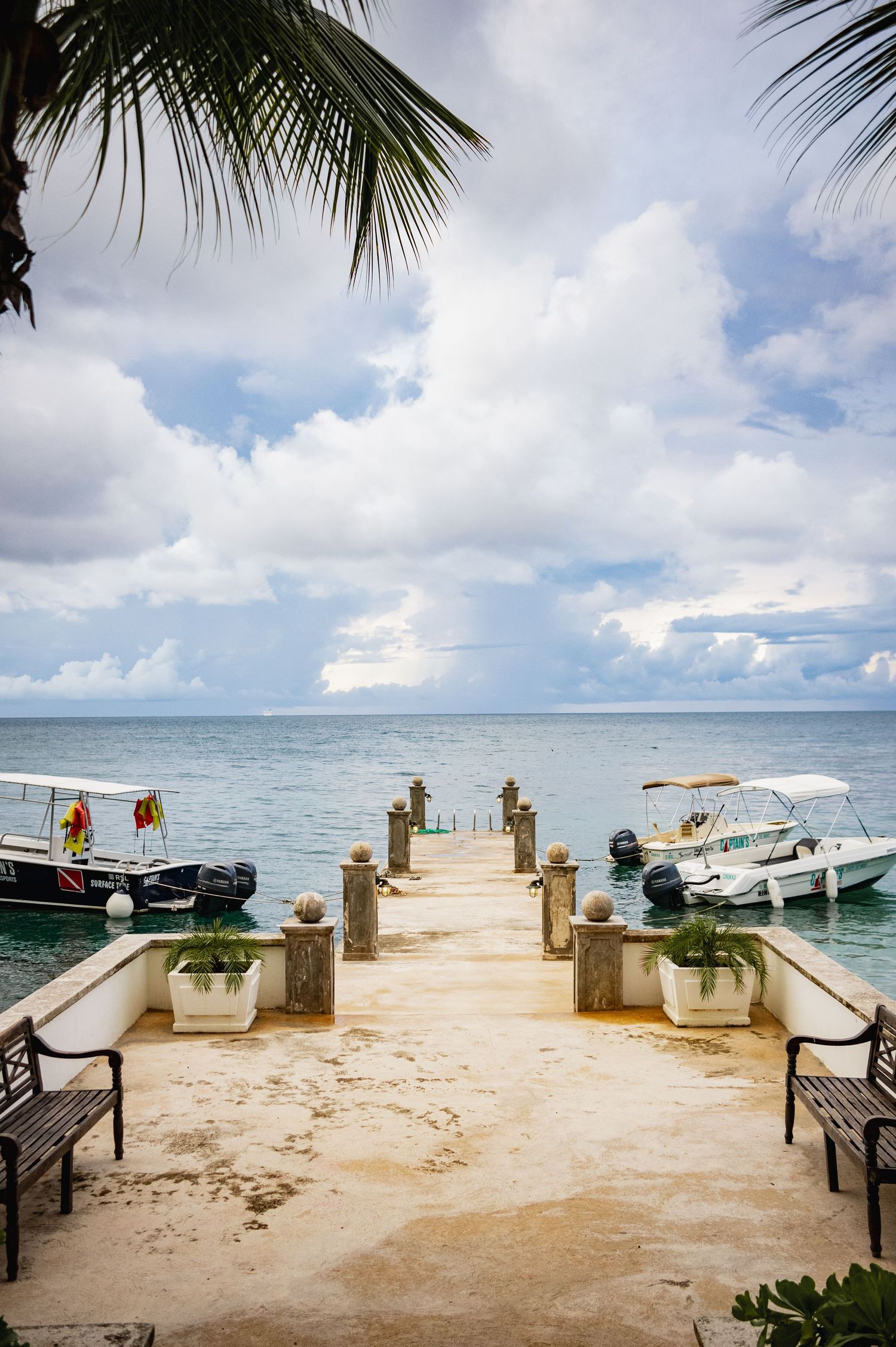 A panoramic view of a wooden pier with benches and boats on a tropical beach under cloudy blue skies.
