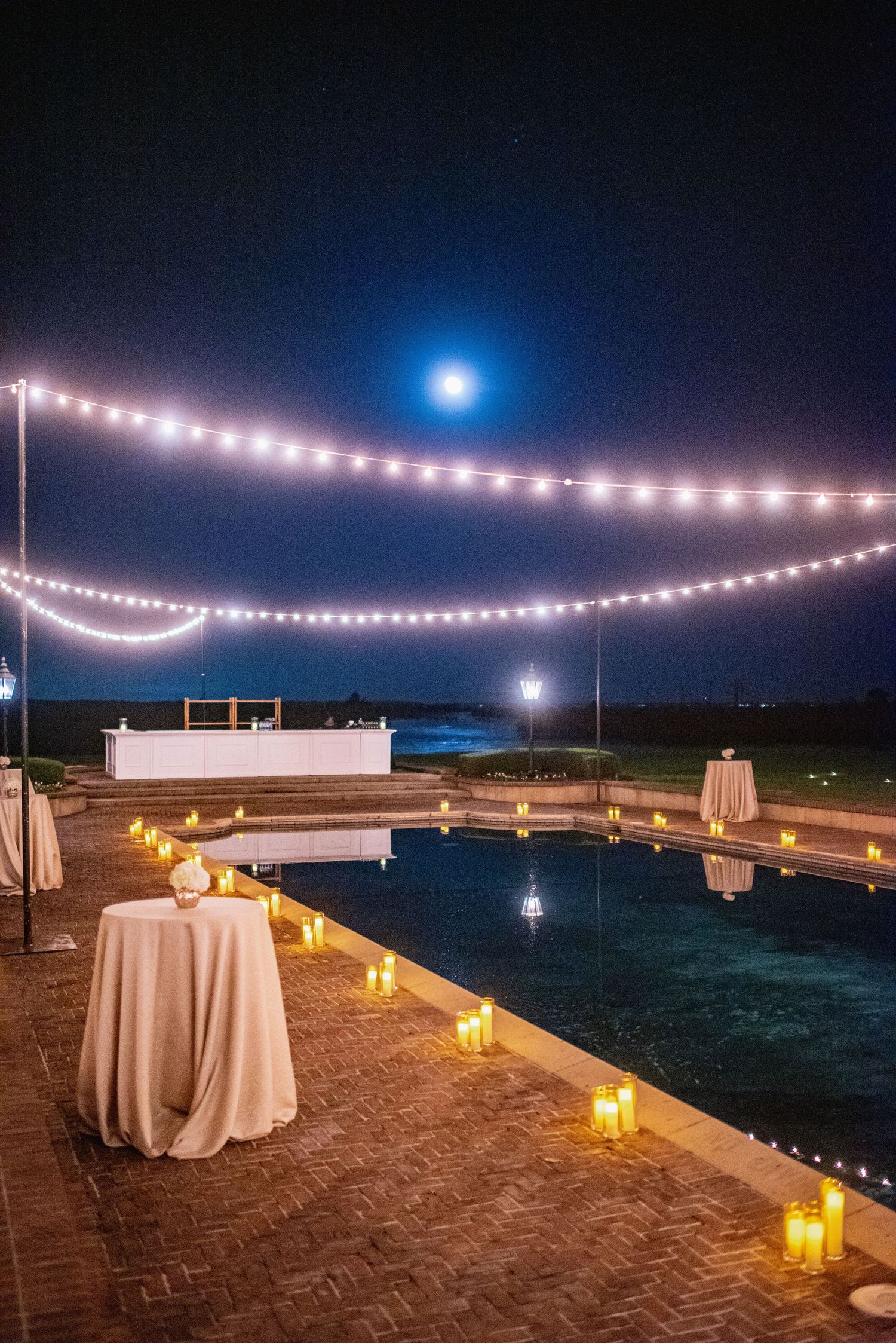 String lights and glowing lanterns illuminate an elegant outdoor event setup along a beachfront at dusk.