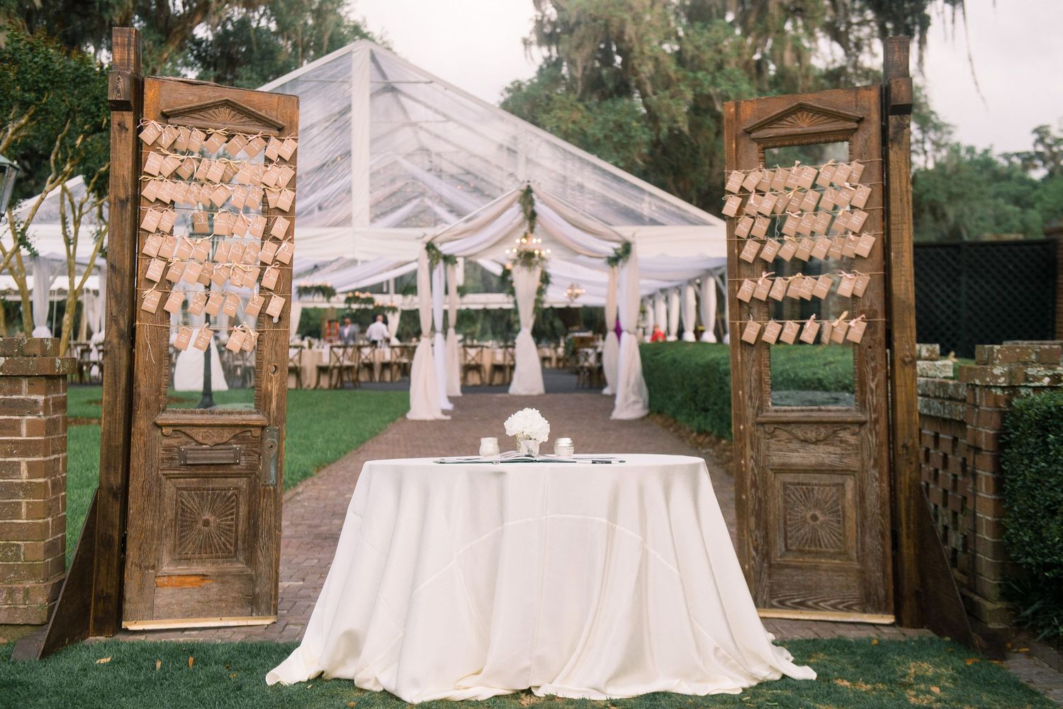 Elegant white wedding tent reception setup with brick entrance columns and natural outdoor surroundings.