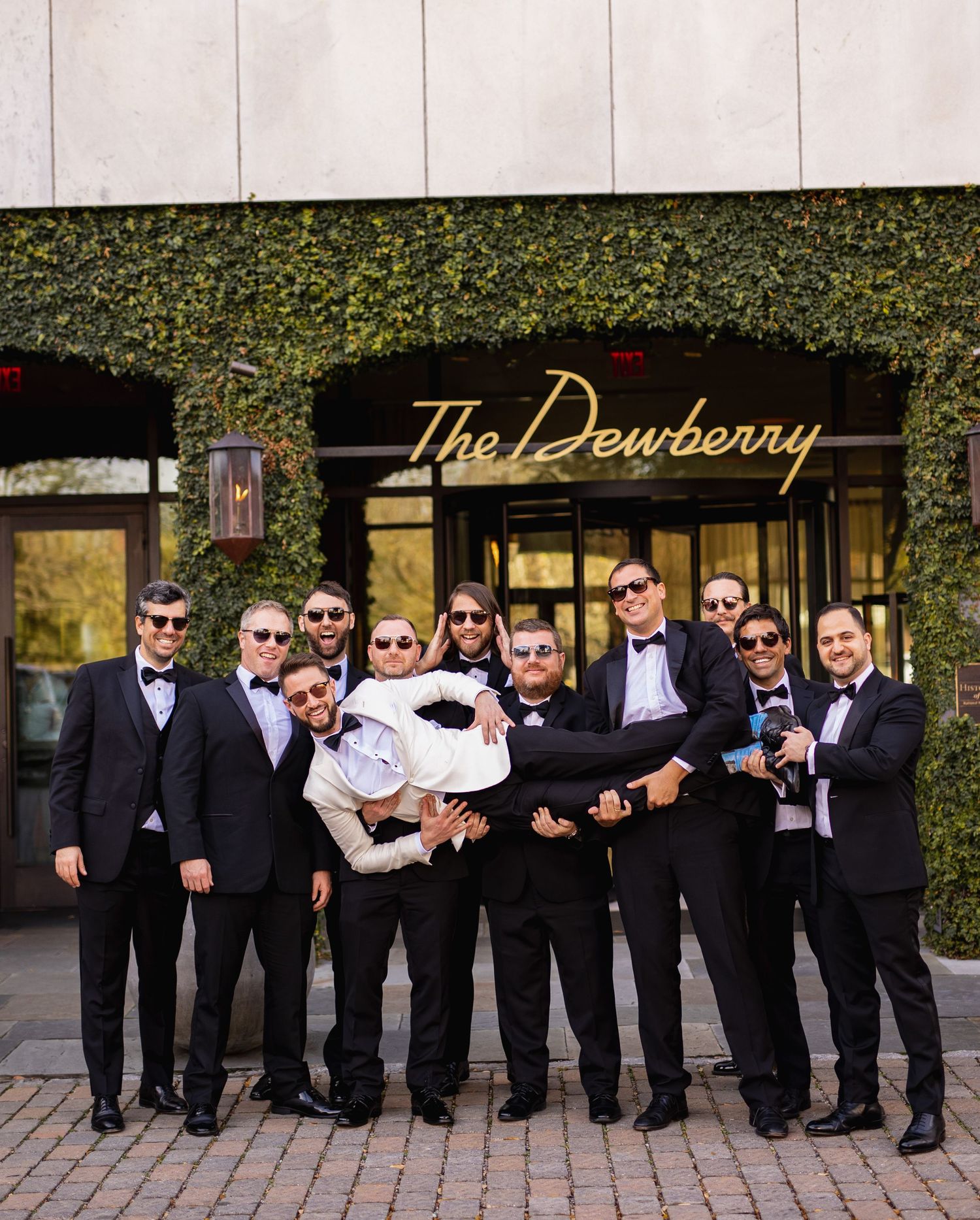 Groomsmen in black tuxedos and bowties strike playful poses in a line against a stone wall backdrop.