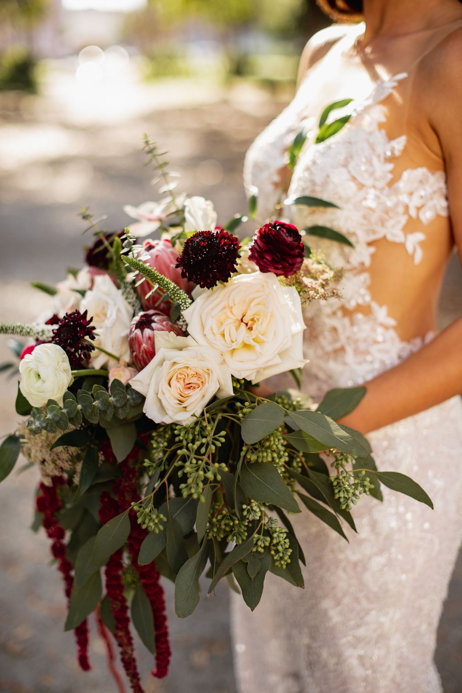 A romantic wedding bouquet features cream roses, deep burgundy dahlias, and cascading greenery against a lace dress.