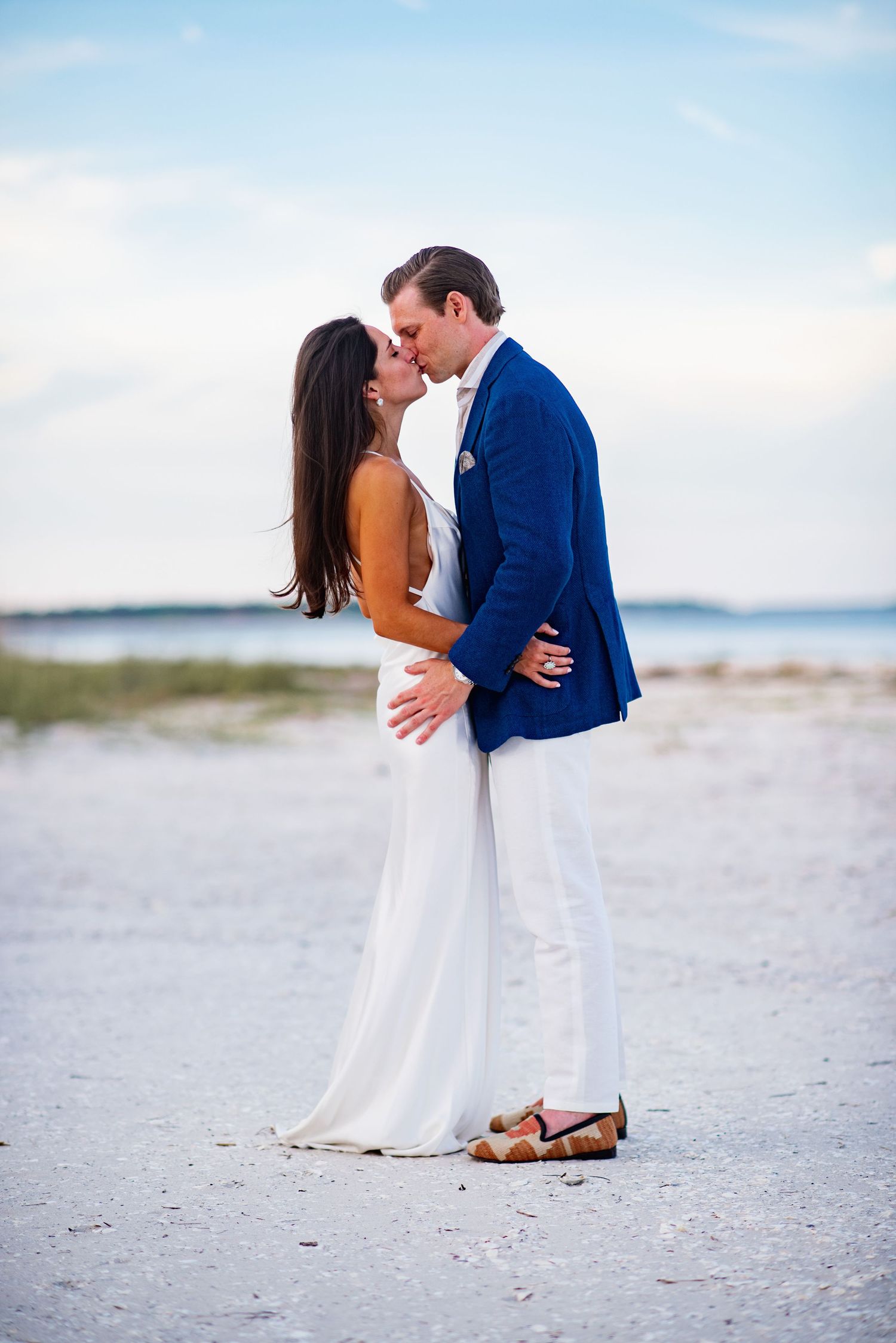 A couple shares a romantic kiss on a white sandy beach with ocean views in the background.
