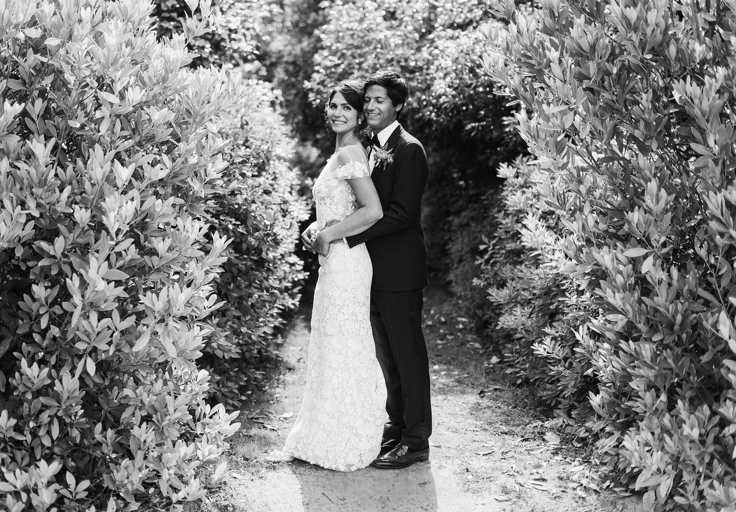A romantic wedding couple embraces in a lush garden pathway creating an elegant black and white portrait.