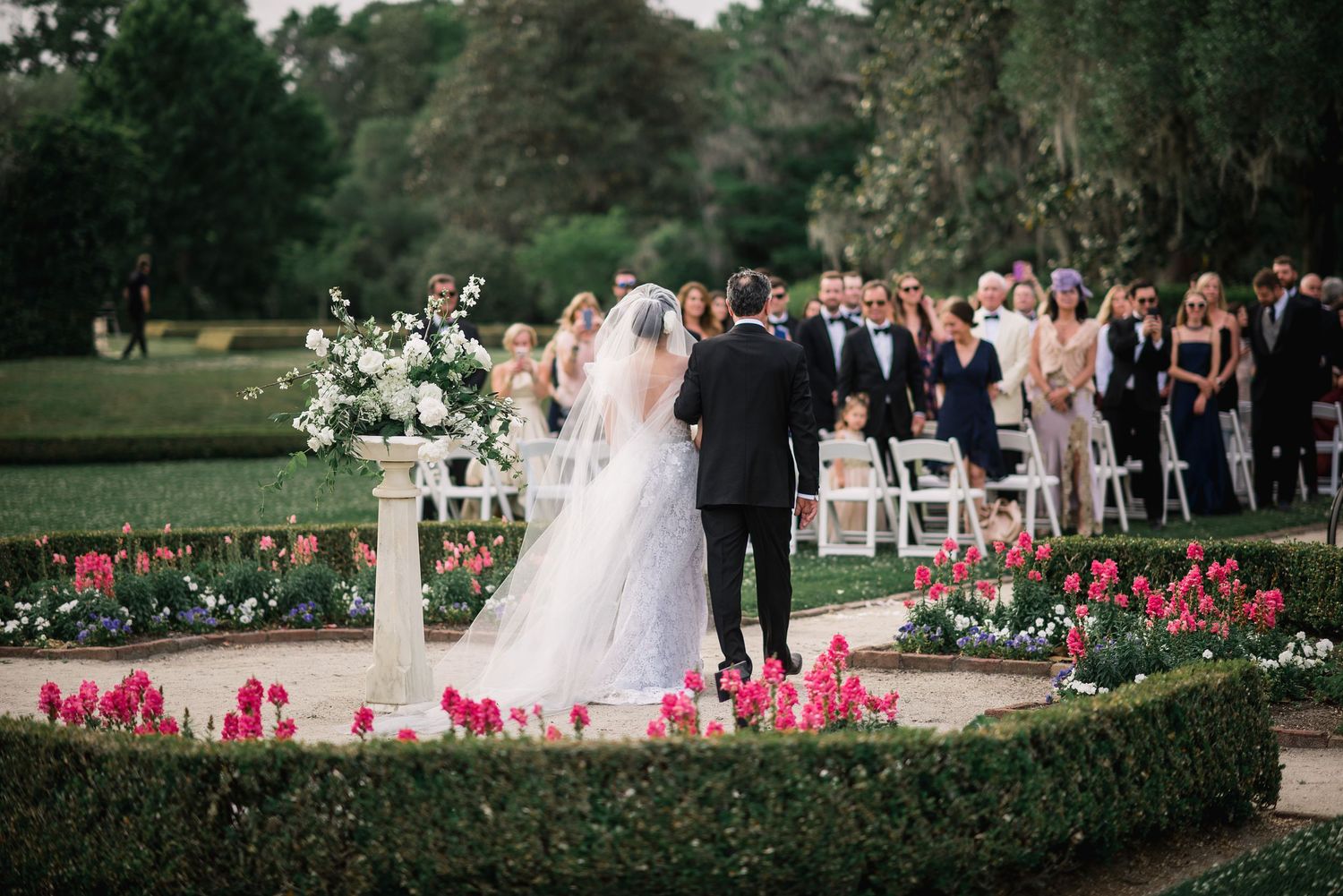 Outdoor garden wedding ceremony setup with pink floral arrangements and white chairs along a stone pathway.
