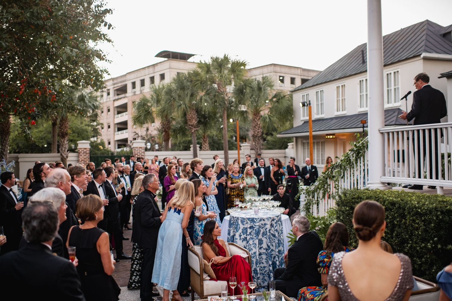 Wedding guests gather outdoors at a coastal venue during sunset celebration with palm trees and buildings in background.
