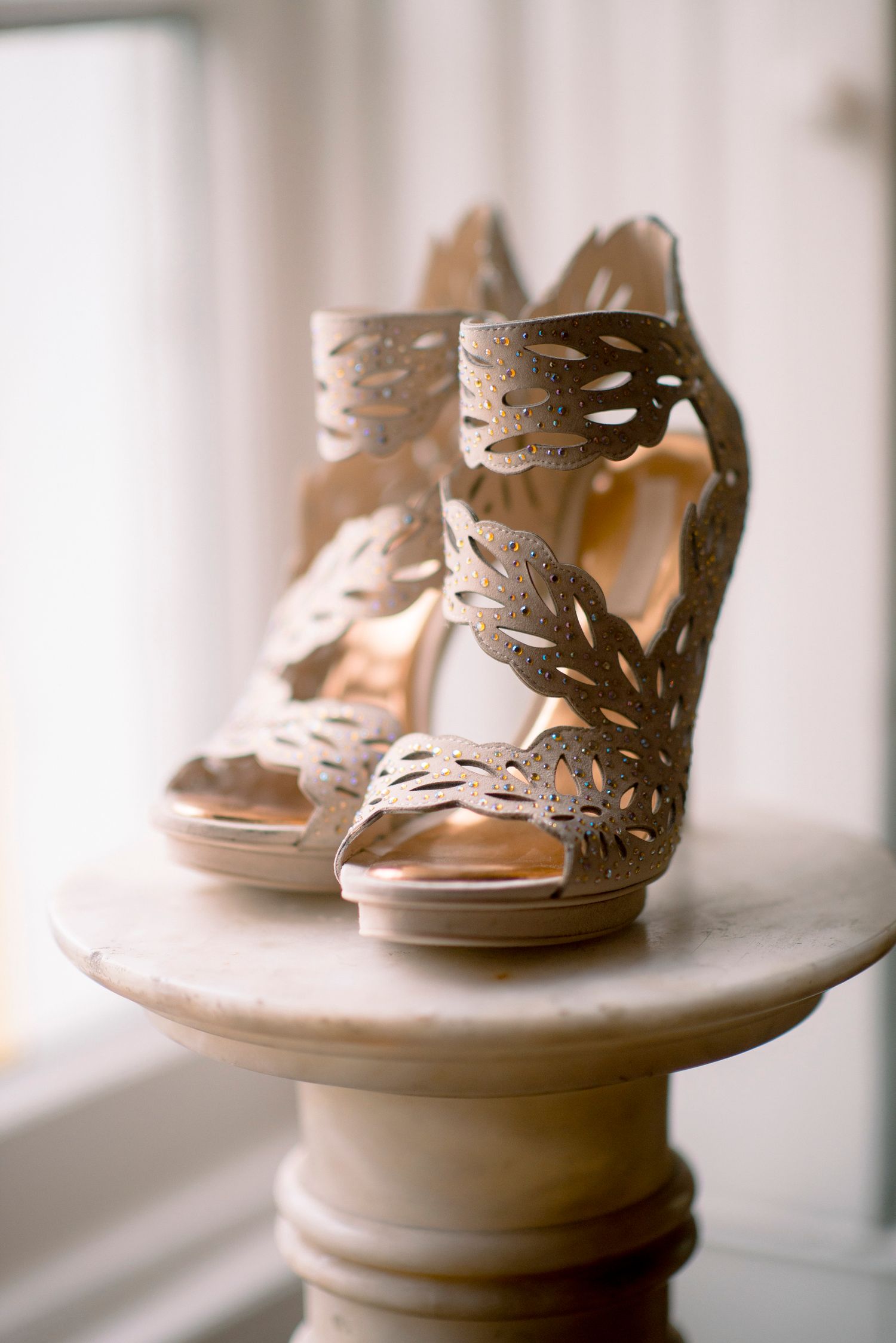 Elegant nude leather sandals with intricate leaf cutout details displayed on a white pedestal table near a window.