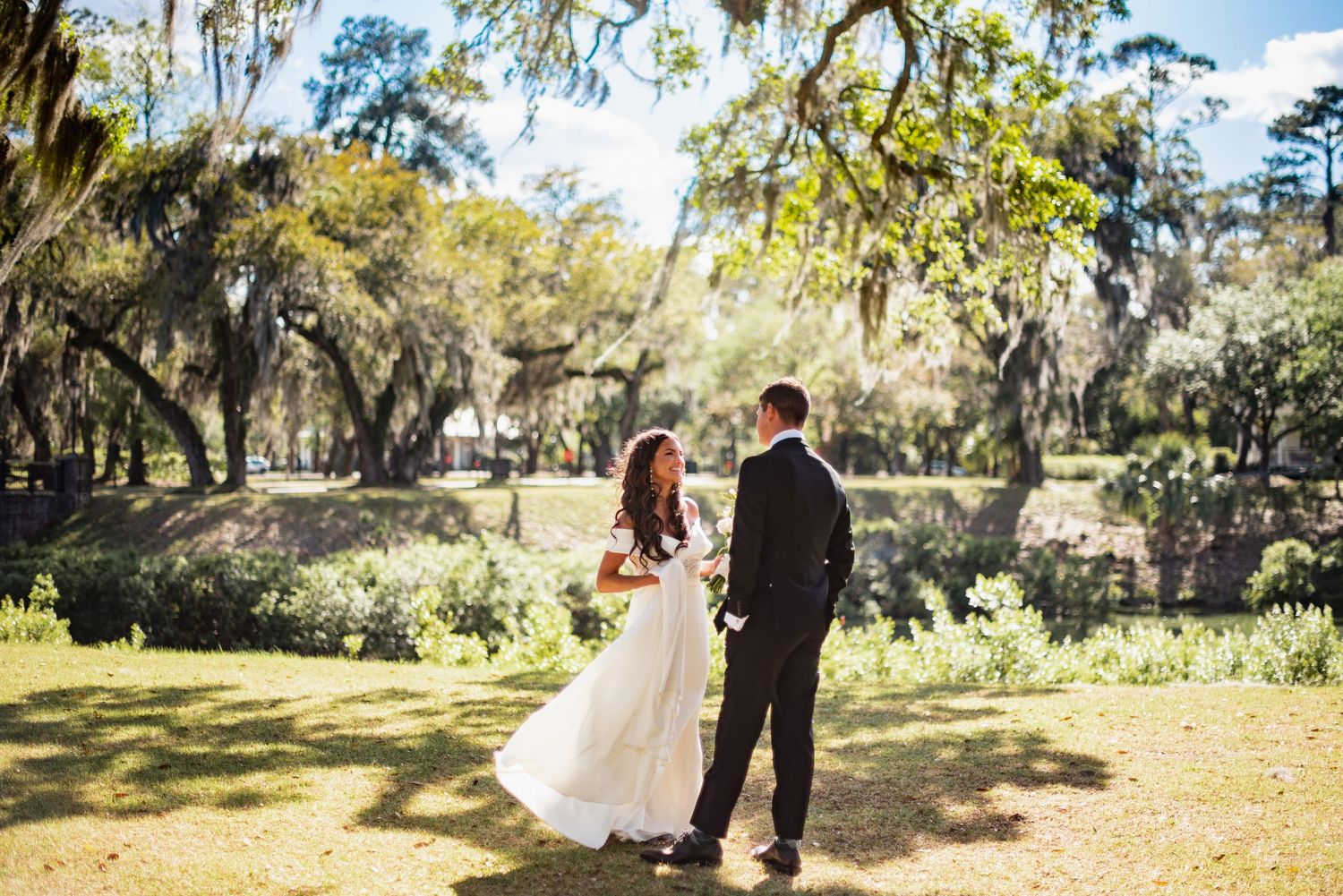 A couple in formal wedding attire walks together on a scenic outdoor path surrounded by Spanish moss and trees.