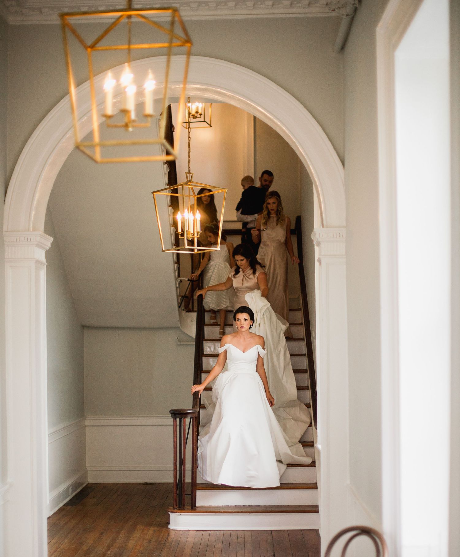 A bride in a white ballgown descends an elegant staircase in a bright interior space with arched doorways and chandeliers.