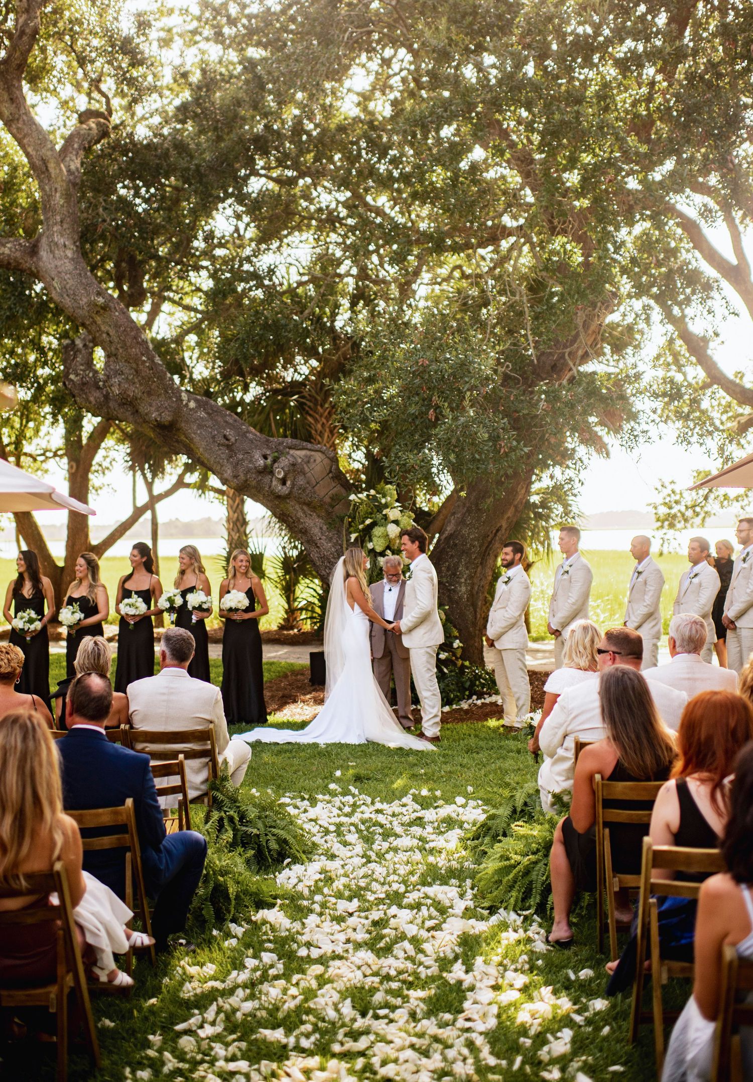 Outdoor garden wedding ceremony with wooden chairs surrounded by blooming white daisies and shaded by mature trees.