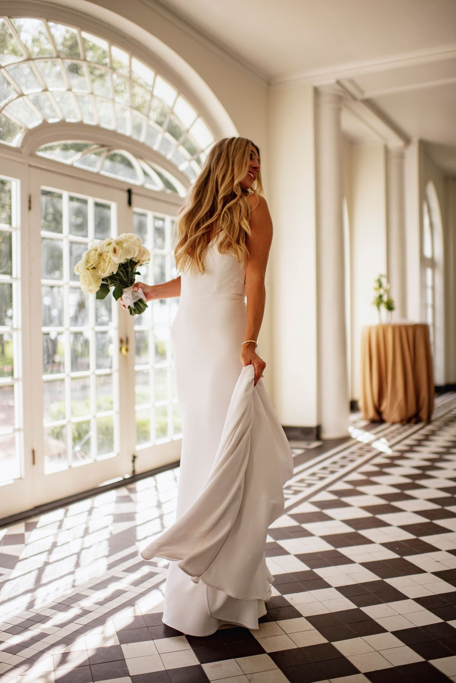 A figure in a flowing white dress twirls and dances through a sunlit hallway with black and white checkered floors.