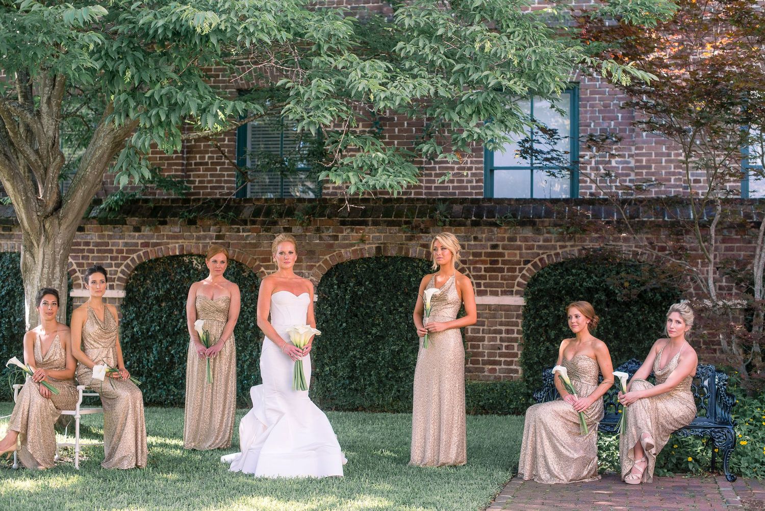 A wedding party poses in front of a brick building with lush greenery, wearing elegant champagne-colored gowns.