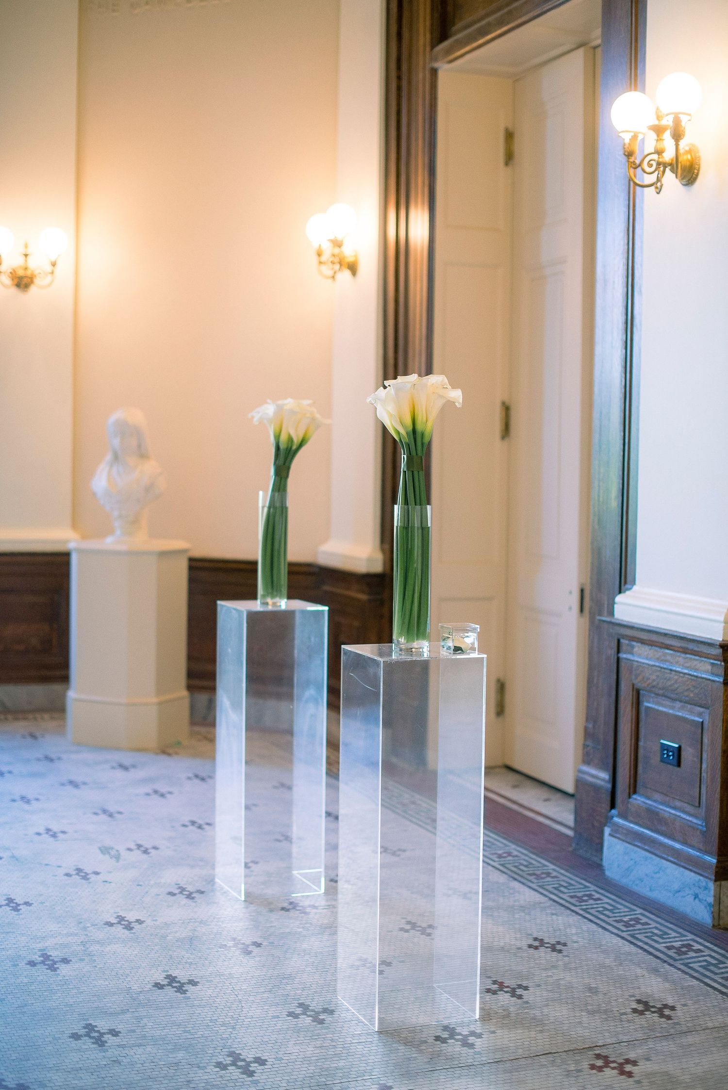 Elegant hotel hallway with white walls, wall sconces, and tall glass vases filled with green stems on marble floor.