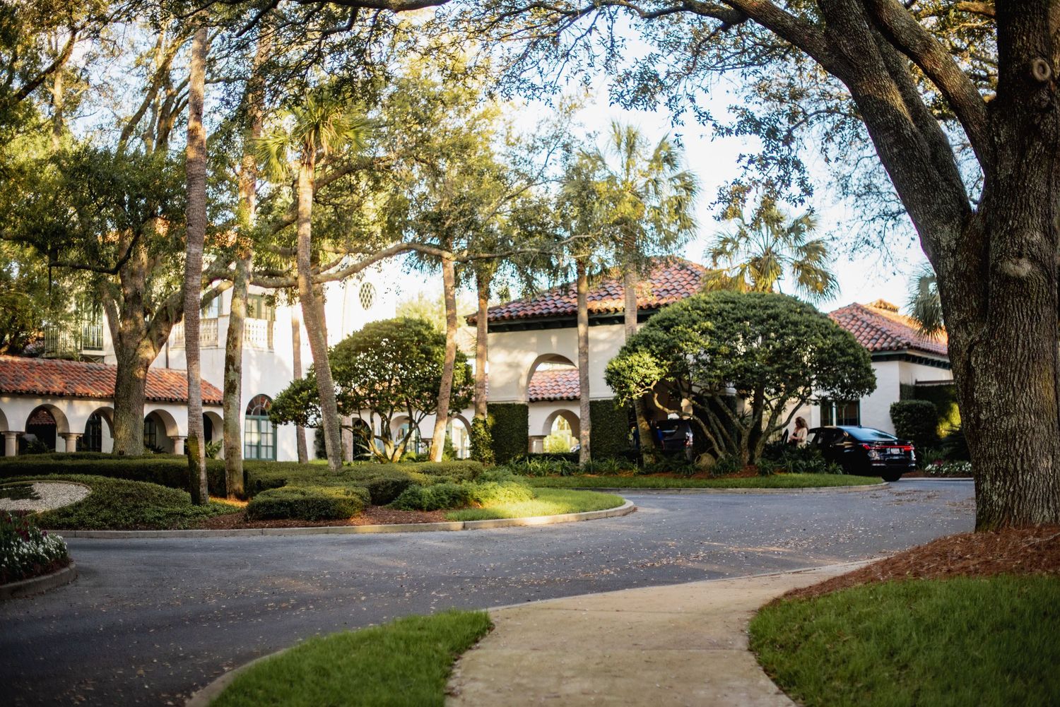 A panoramic view of a tree-lined driveway leading to a Mediterranean-style building with arched walkways.