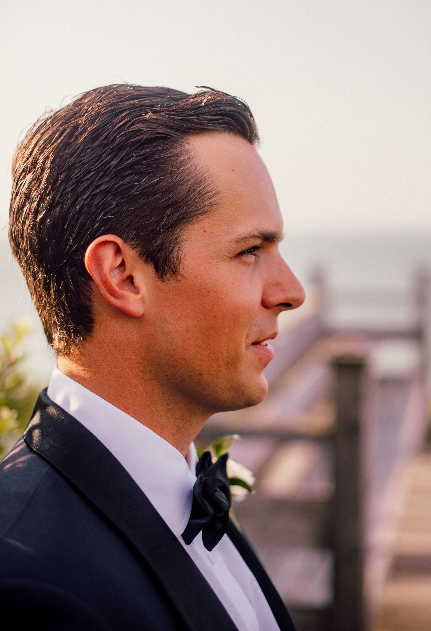 Profile view of groom in black tuxedo and bowtie at sunset.