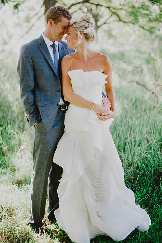 A romantic outdoor wedding photo showing a couple in a gray suit and white gown embracing in a sunlit natural setting.