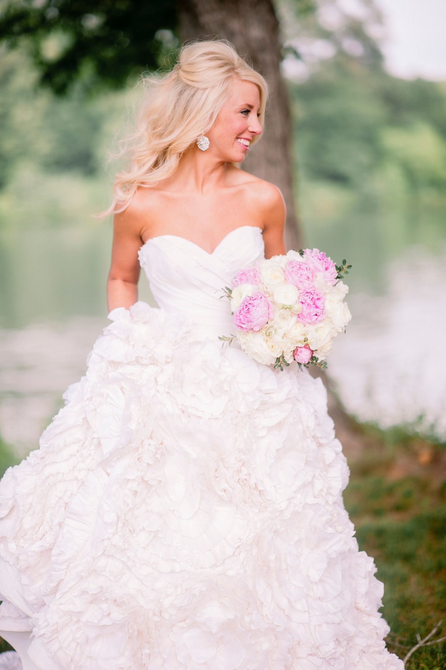 Bride in luxurious ruffled ballgown poses with pink peonies in an outdoor garden setting.