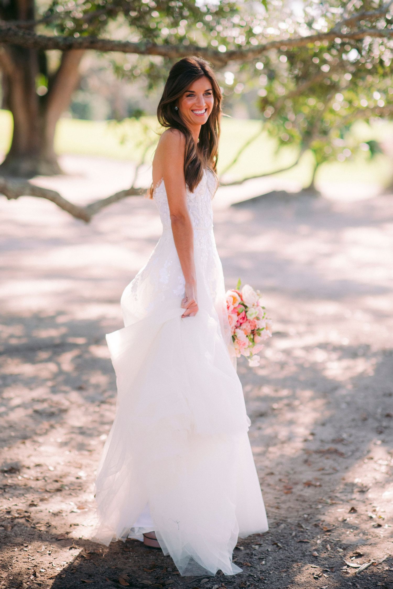 Bride twirling in flowing white gown under sunlit trees.