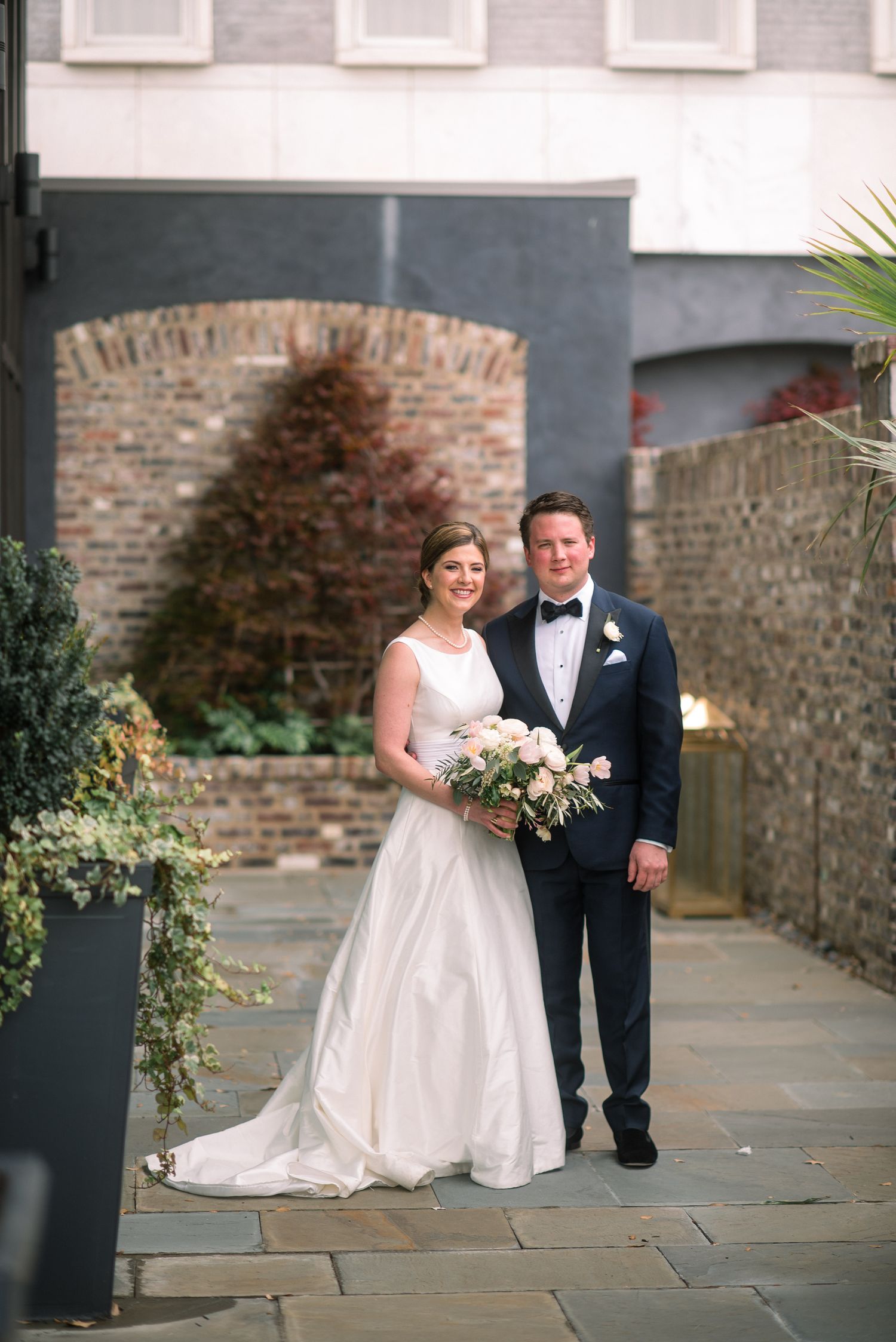 A bride and groom walk together along a stone pathway beside a brick wall during their wedding photoshoot.