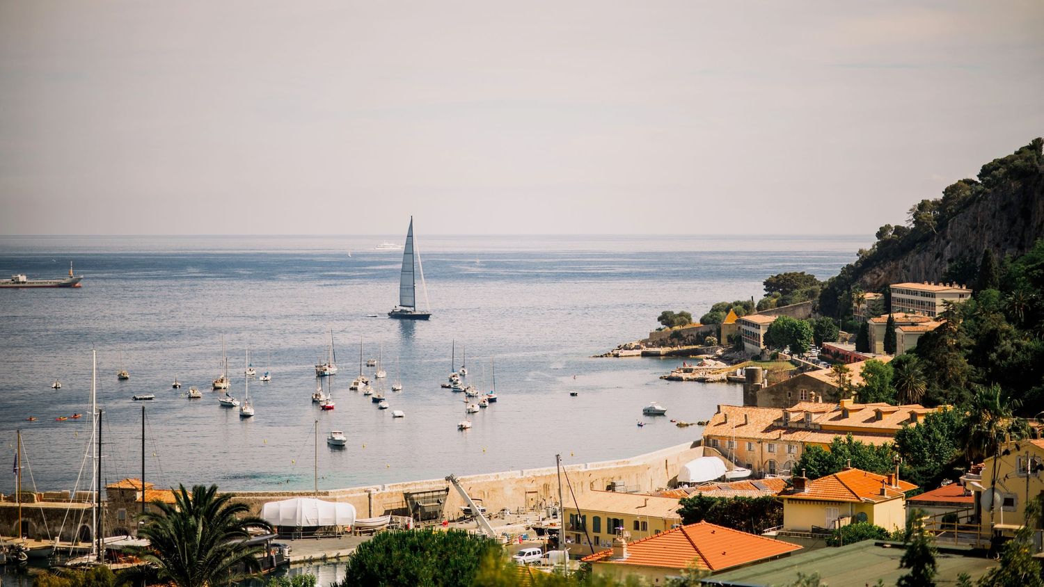 Coastal panorama showing sailboats anchored in a bay with terracotta roofed buildings on shore.