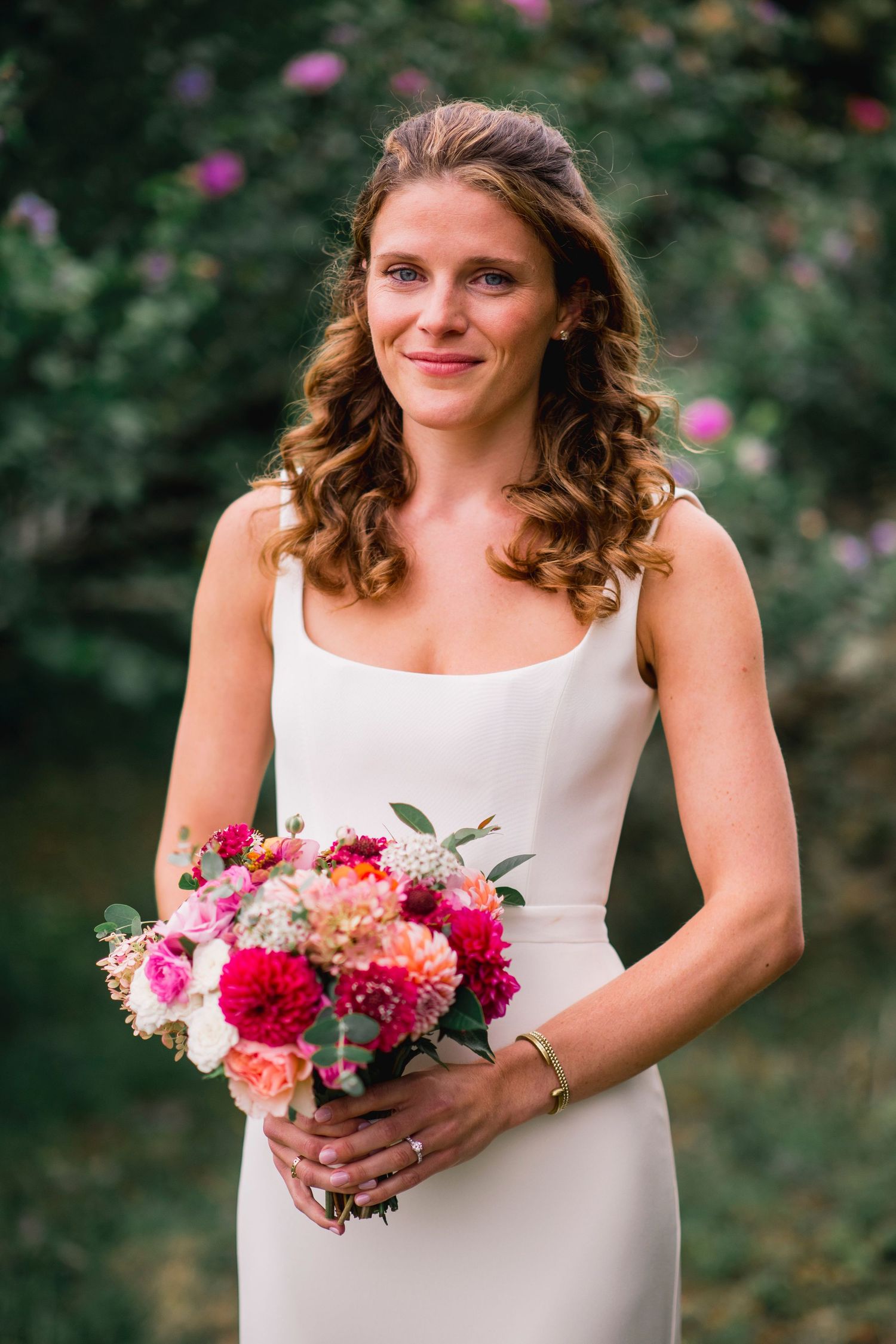 Person in white dress holds colorful bouquet of pink and coral flowers in garden setting.